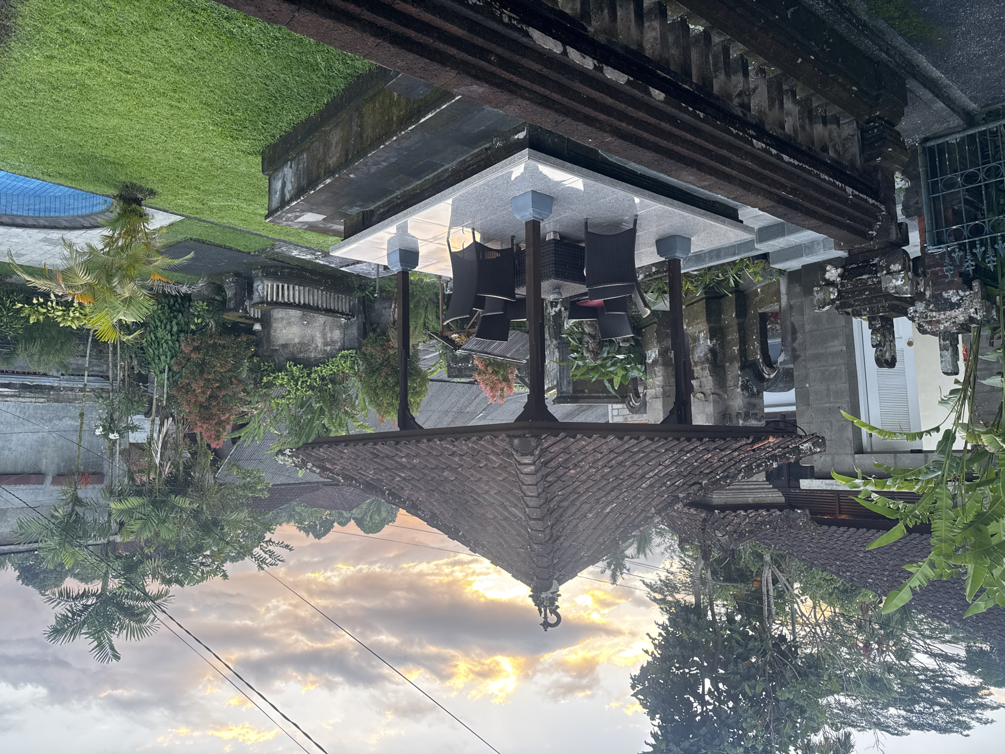 The image depicts a scene in Ubud District, Indonesia, showcasing a traditional Balinese residence at dusk. The foreground features a tiled roof with intricate carvings, extending into the frame. Underneath, a raised, open pavilion with a modern grey tiled top, supported by dark-colored pillars, hosts a seating area with dark wicker chairs and a table. Lush greenery and trees surround the structures, with a view towards the horizon where the sky exhibits a sunset gradient of pale yellow and grey clouds. Power lines traverse the sky. A swimming pool is visible in the background, surrounded by a well-maintained lawn and traditional Balinese architectural elements. The overall scene suggests a blend of traditional Balinese architecture and modern amenities in a serene outdoor setting.