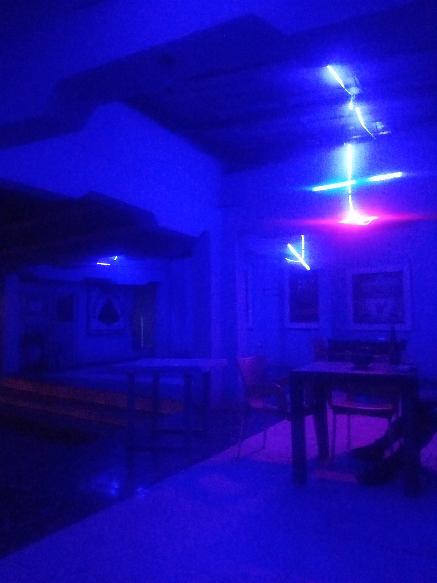 A dimly lit interior space, possibly a restaurant or bar, is illuminated by bright blue and pink neon lights. Two tables with chairs are visible, one in the foreground with a dark tabletop, and another further back with a lighter surface. On the back table, a dark object, potentially a bottle, sits near two framed pictures on the wall. The walls appear to be painted a light color, contrasting with the intense lighting. Shadows are cast across the floor and walls, creating a dramatic and somewhat abstract atmosphere. The overall impression is one of a vibrant and modern establishment.