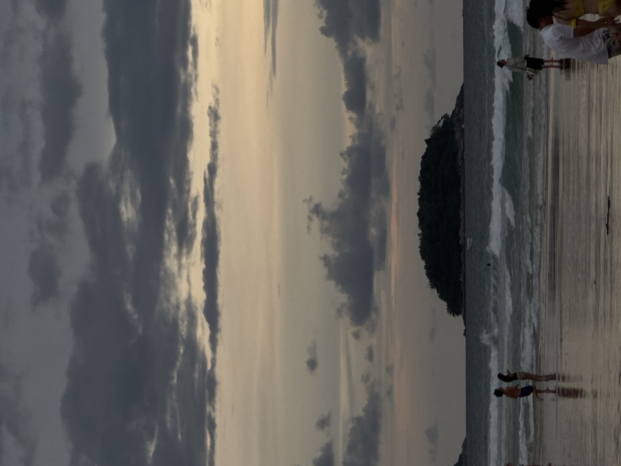 The image depicts a beach scene in Karon, Thailand, during twilight. The sky is filled with dramatic, dark clouds. In the foreground, the wet sand reflects the subdued light, with several people scattered across it. To the left, a man and woman stand facing the water. Further to the right, a woman stands in the shallow water, holding an object. Towards the far right, a couple stands close together. In the middle ground, the ocean churns with moderate waves, and a small, tree-covered island is visible in the distance.