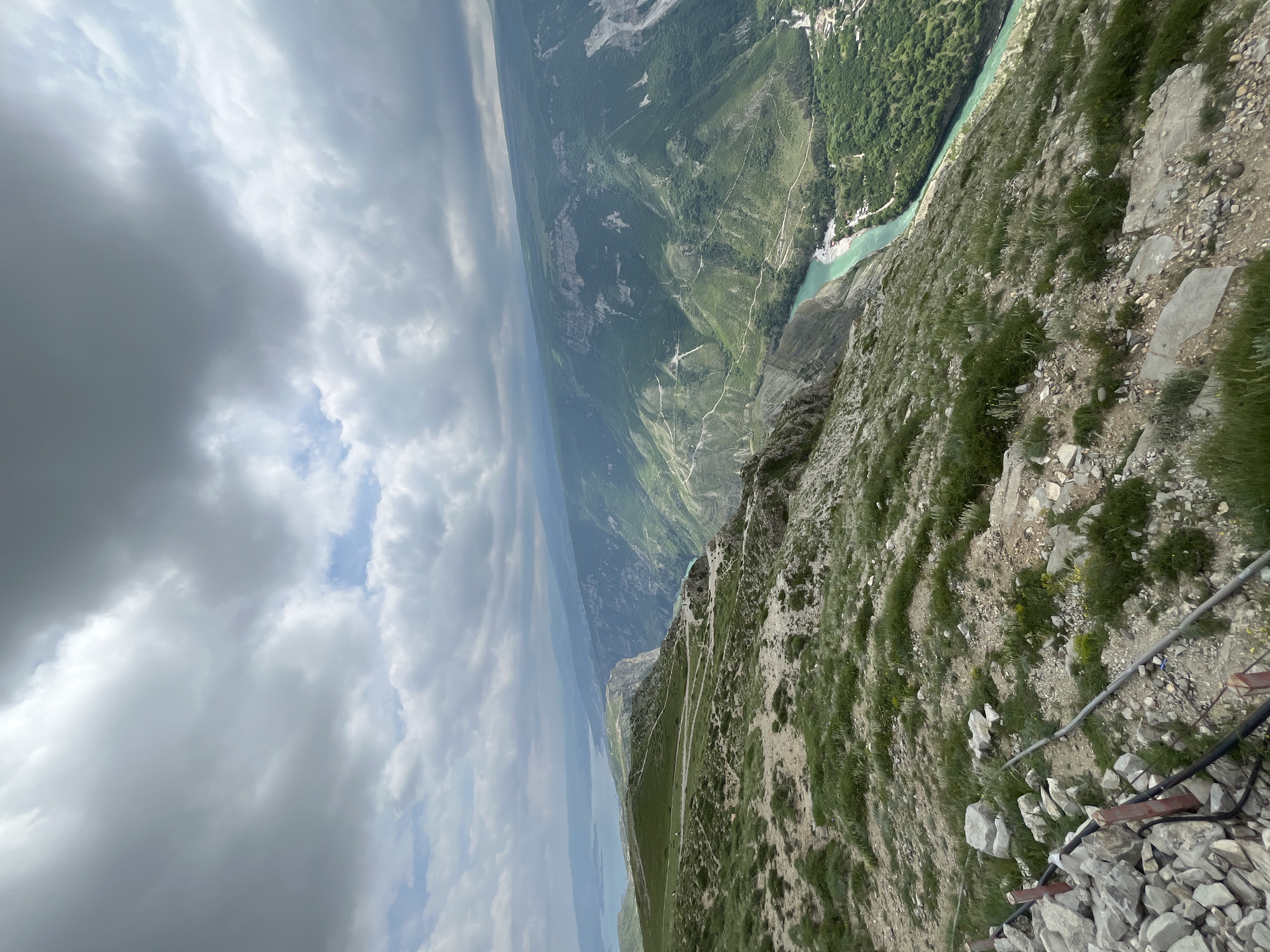 An elevated, angled landscape photograph focuses on a steep, grassy mountainside descending into a river valley. The sky is overcast with heavy, gray clouds, allowing glimpses of blue in patches. In the distance, a wide river flows through the valley, bordered by dense green forests and terraced slopes. The mountainside in the foreground is covered with sparse grass and rocky terrain, with exposed cables and metal supports near the bottom of the frame. The location is near the city of Dubki, Russia.