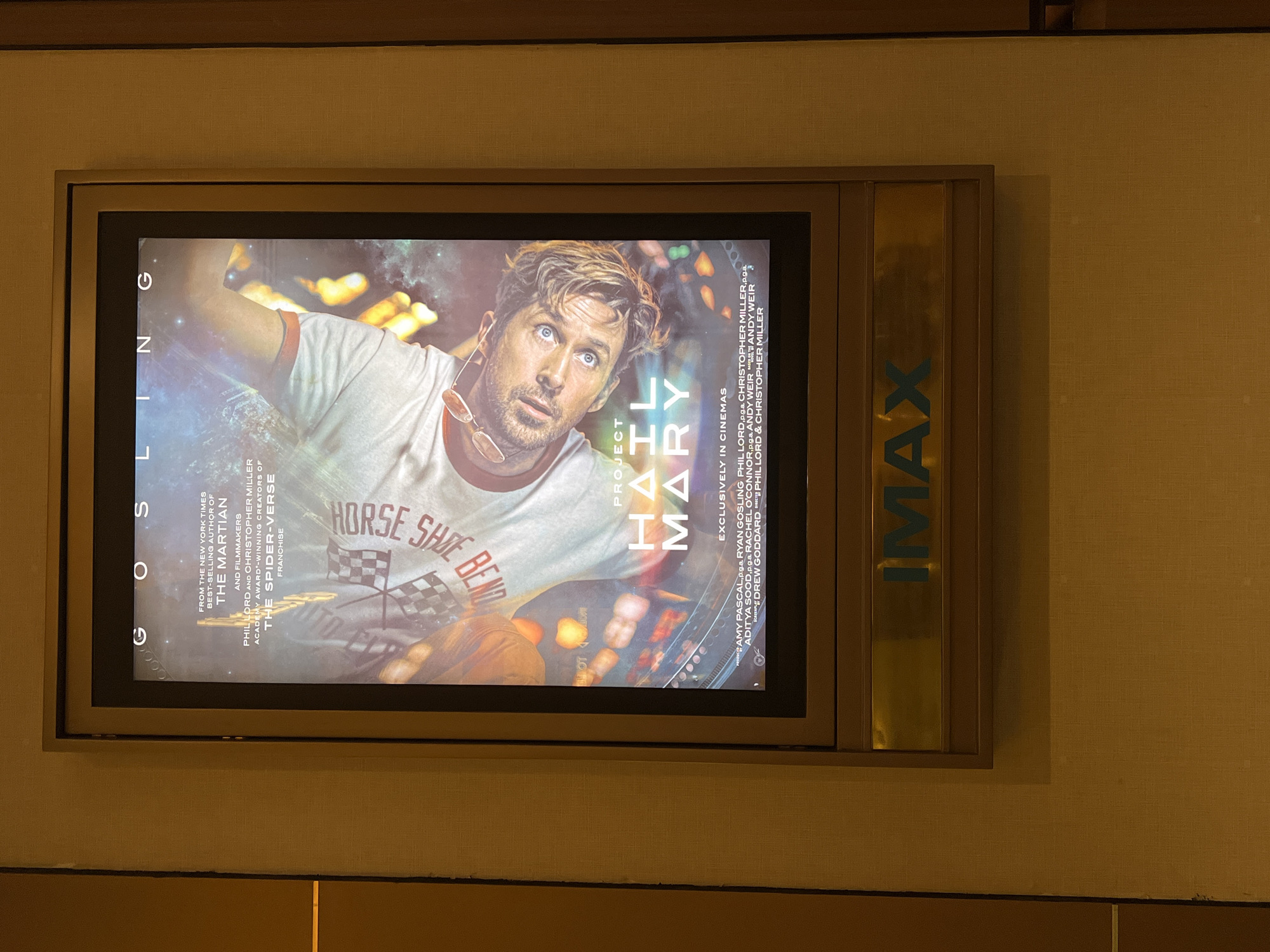 The image shows a promotional poster for the movie "Project Hail Mary," starring Ryan Gosling. The poster is displayed in a backlit frame with an "IMAX" logo on the right side. The poster promotes Ryan Gosling, the filmmakers Phil Lord and Christopher Miller (of The Martian and Spider-Verse fame), and indicates the film is exclusively showing in cinemas. The poster is mounted on a textured, neutral-colored wall. Based on the visual evidence in the photo alone, I cannot confirm this is in Denpasar, Indonesia.
