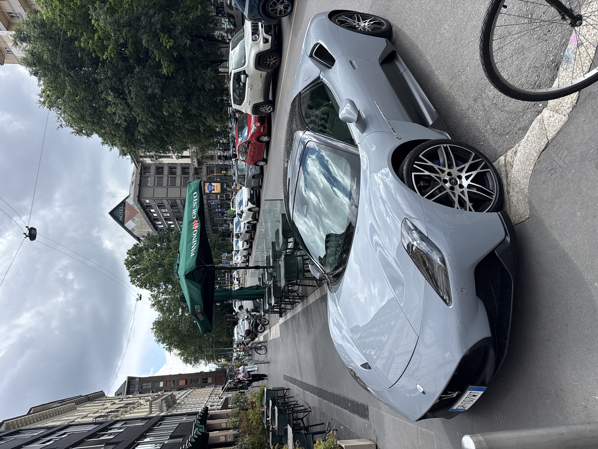 The image shows a gray Maserati MC20 parked on a street in Milan, Italy. The car is positioned on the right side of the frame, with the front of the car facing towards the foreground. It has black rims with silver accents. The license plate reads "GP 107HT". Behind the car, a row of parked vehicles is visible, including a white Jeep and a red Fiat. On the left side of the frame, there is a sidewalk with outdoor seating under green "PANINO GIUSTO" branded umbrellas. Buildings line the street on both sides. A bicycle is parked near the car on the right. People can be seen walking in the distance. An overhead wire extends above the umbrellas and seating area. The sky is overcast.