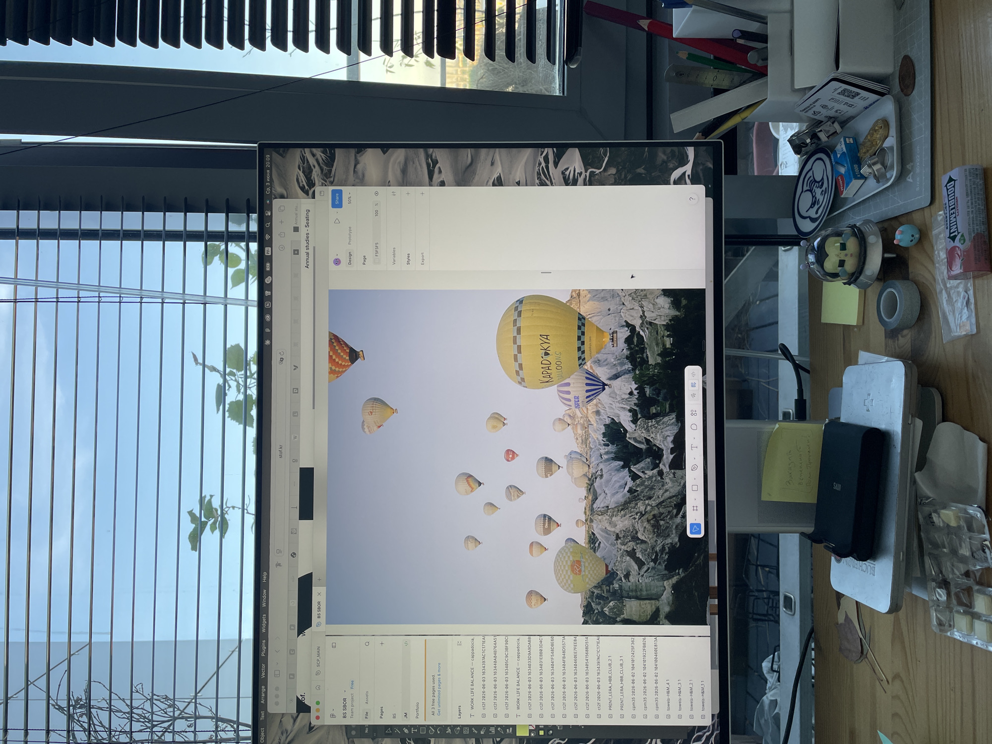 The image displays a workspace featuring a computer monitor, an adjacent window, and various desk items. The monitor is primarily showing design software, likely Figma, with a large image of numerous hot air balloons floating above a mountainous, rocky landscape. Text visible on one yellow balloon clearly states "KAPADOKYA BALLOONS," indicating the scene is set in Cappadocia, Turkey. Another balloon has "SFER" visible.
The software interface on the monitor shows a project titled "Annual studies - Seating." On the left panel, sections like "Layers," "Pages," and "Assets" are visible, with project names including "BS SBOR," "SCP_MAIN," and "WORK LIFE BALANCE - cappadocia." A tab labeled "stof.kr" is also open. The right panel includes options for "Design" and "Prototype," scaling ("50%", "100%"), and sections for "Variables," "Styles," and "Export." The top bar of the monitor shows "Ср, 3 июня 20:09," which translates to "Wednesday, June 3, 8:09 PM" in Cyrillic, suggesting the time the photo was taken.
To the right of the monitor, a collection of desk items is arranged on a light wooden surface. These include a stationery holder containing pencils and a ruler, a small white tray with various metallic trinkets and a circular blue and white logo-emblazoned disc (resembling the GE logo), and a small white box labeled "x1000." Further right, there is a clear dome-shaped figurine, a small light blue blob-shaped figure, a grey tape roll, and a black device (possibly a power bank) labeled "SAM." A yellow sticky note with handwritten Cyrillic text, including "Заказать" (Order) and "Вернемся" (We'll return), is attached to a white box that has "BUCHENOM" printed on it. A cutting mat with "A4 Size 30x22 cm" and measurement markings is also visible, next to a white and red box for "DOUBLMINT Chewy Mints" and a transparent plastic box with compartments.
To the left of the monitor, a window with partially open horizontal blinds reveals a bright sky outside and some green foliage. Given the Cyrillic text on the monitor and the sticky note, the location context provided (Unknown, Russia) is consistent with the visible linguistic cues.