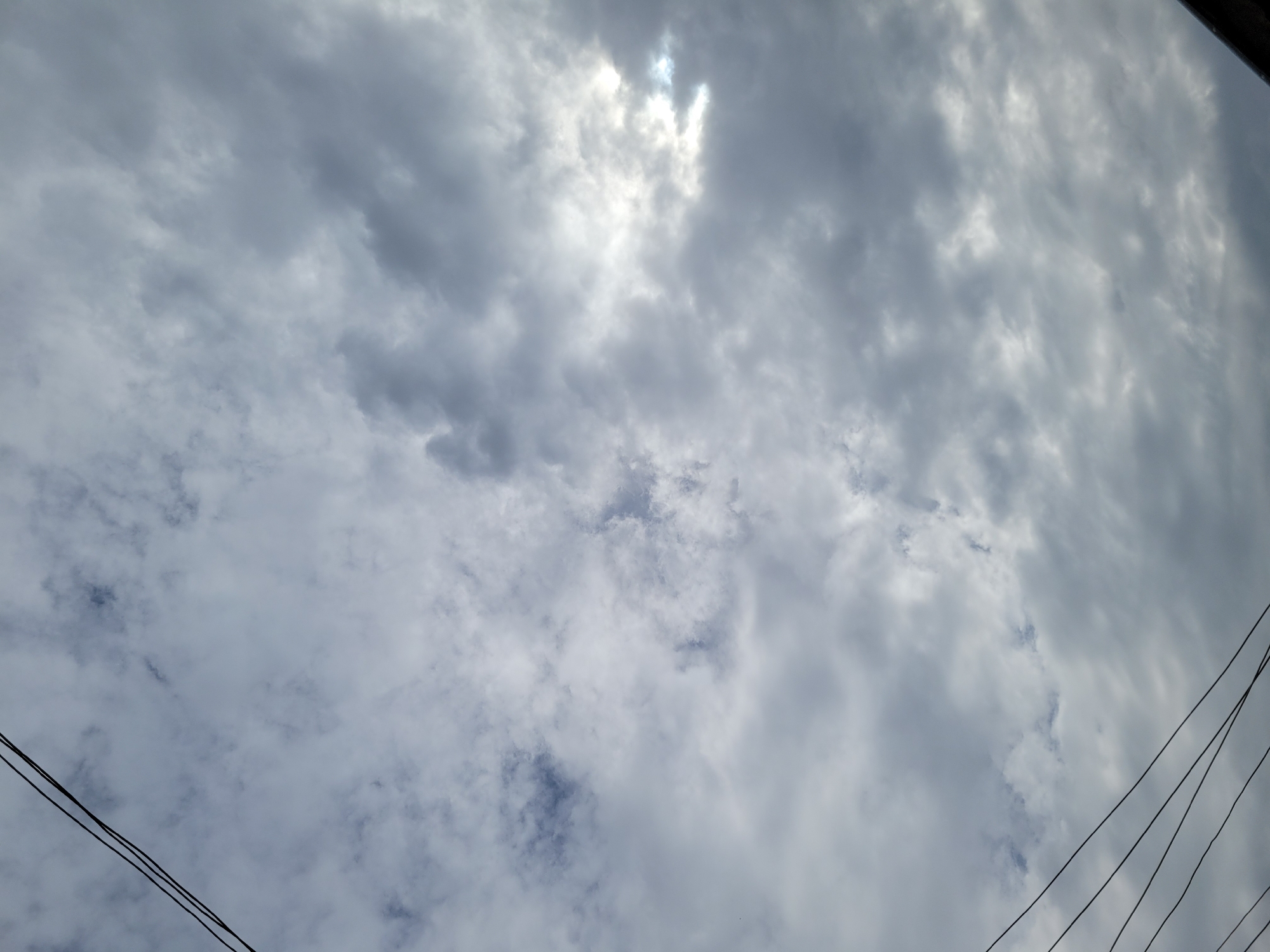 The image depicts a cloudy sky with power lines in the foreground. The clouds are a mix of grey and white, suggesting a partly cloudy or overcast day. The sun is not directly visible but is partially obscured by a bright patch of clouds, indicating a possible break in the cloud cover. The power lines, appearing black and thin, are diagonally oriented across the frame, stretching from the top left to the bottom right. The scene appears to be an outdoor environment, likely within a city setting due to the presence of power lines. Without additional information or context, the specific location within Jos, Nigeria, cannot be determined from the image alone. There are no people, animals, or specific structures visible that would offer further clues about the exact location or activity. The overall impression is a typical sky view, common in urban areas.