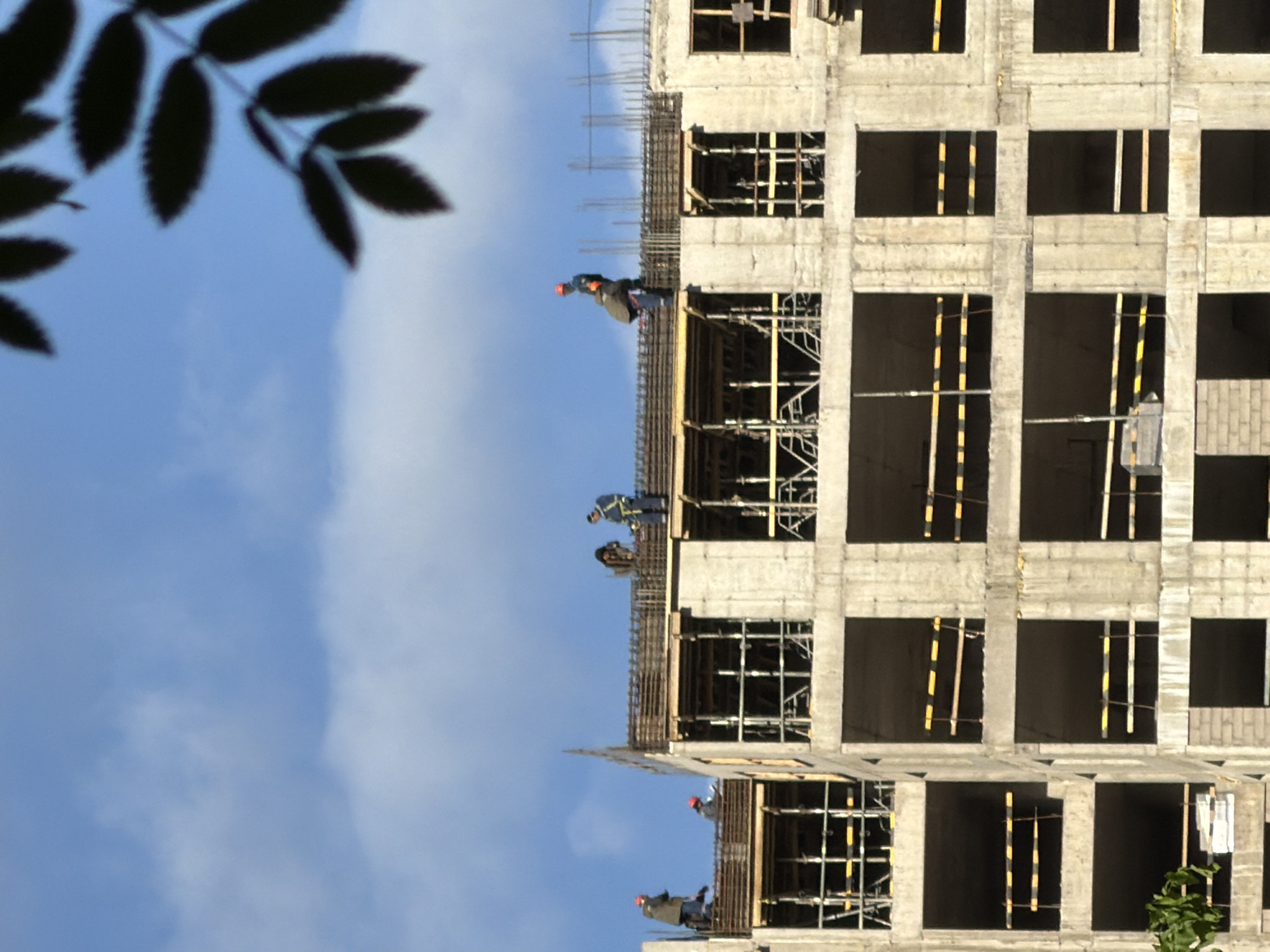The image shows a building under construction in Saint Petersburg, Russia. The building has multiple floors with open window spaces and scaffolding. Several construction workers are visible on different floors, engaged in their tasks. The sky is visible in the background, with some foliage in the foreground.