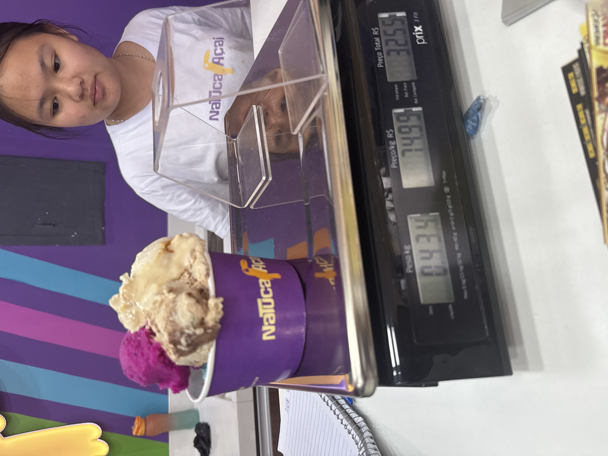 The image depicts an indoor scene, likely a retail establishment in Cabo Frio, Brazil, given the "Naluca Açaí" branding on the product cup and the employee's shirt. A young woman, likely an employee, is partially visible in the background, wearing a white t-shirt with the "Naluca Açaí" logo. She is looking down and to the left, with a clear plastic guard between her and the foreground.
In the foreground, a digital scale is prominent. On the scale, a purple paper cup filled with three scoops of what appears to be ice cream or açaí bowl topping sits. The scoops are of different colors: pink, light brown, and a darker brown swirl. The scale displays readings: "Peso kg" (Weight kg) shows "0.434", and to the right, "Preço Total R$" (Total Price R$) shows "32.55". Another reading, "Preço/kg R$" (Price/kg R$), shows "74.99". The scale itself is black with a metallic trim and a digital display panel.
To the left of the scale, a spiral-bound notebook with lined pages is partially visible, with some writing in blue ink. Behind the notebook, a colorful gradient bottle with an orange cap is standing upright. The background features a wall with diagonal stripes of purple and blue, and a yellow cutout shape, possibly a decoration. The overall impression is a close-up view of a point-of-sale transaction for frozen dessert.
