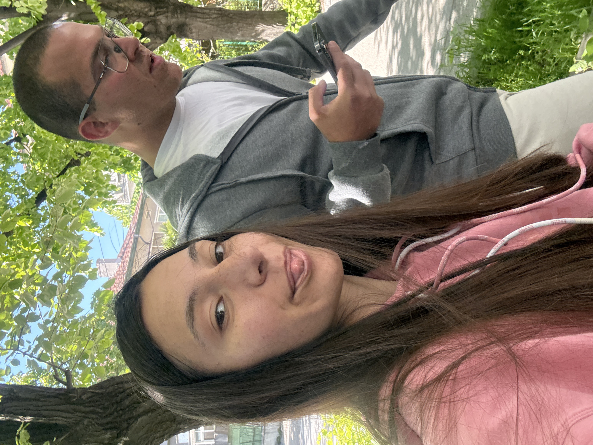 A selfie image shows two individuals in an outdoor urban environment.
One individual is a woman with long, dark hair and a pink hooded sweatshirt. She is closest to the camera. Her expression is playful.
The second individual, a man with short hair and glasses, is wearing a grey hooded sweatshirt over a white t-shirt. He is looking off-camera and appears to be holding a mobile phone.
The background features trees with green foliage. A building can be seen in the distance. Based on architectural styles and common urban features, the location is likely in a city. Although a precise location in Varna, Bulgaria cannot be determined with certainty, the environment is consistent with urban green spaces in the city.