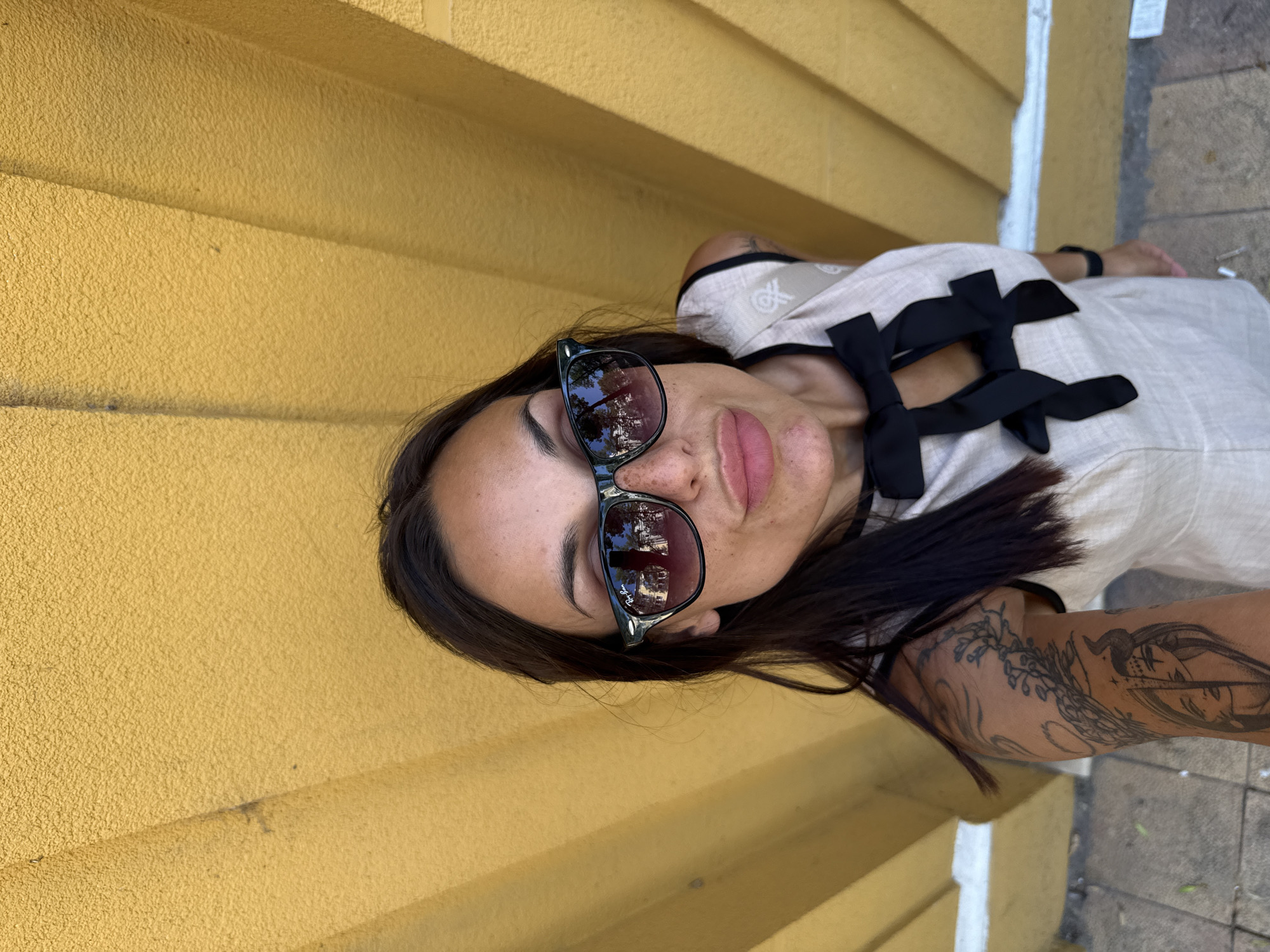 A woman with dark hair, wearing sunglasses and a light-colored dress with black bows, stands in front of a textured yellow wall. Her left arm, partially visible, displays tattoos. The image appears to be a selfie, with the woman's face prominently featured. The backdrop suggests an outdoor urban setting. The specific location in Pleven, Bulgaria is not discernible from the visual information provided in the image.
