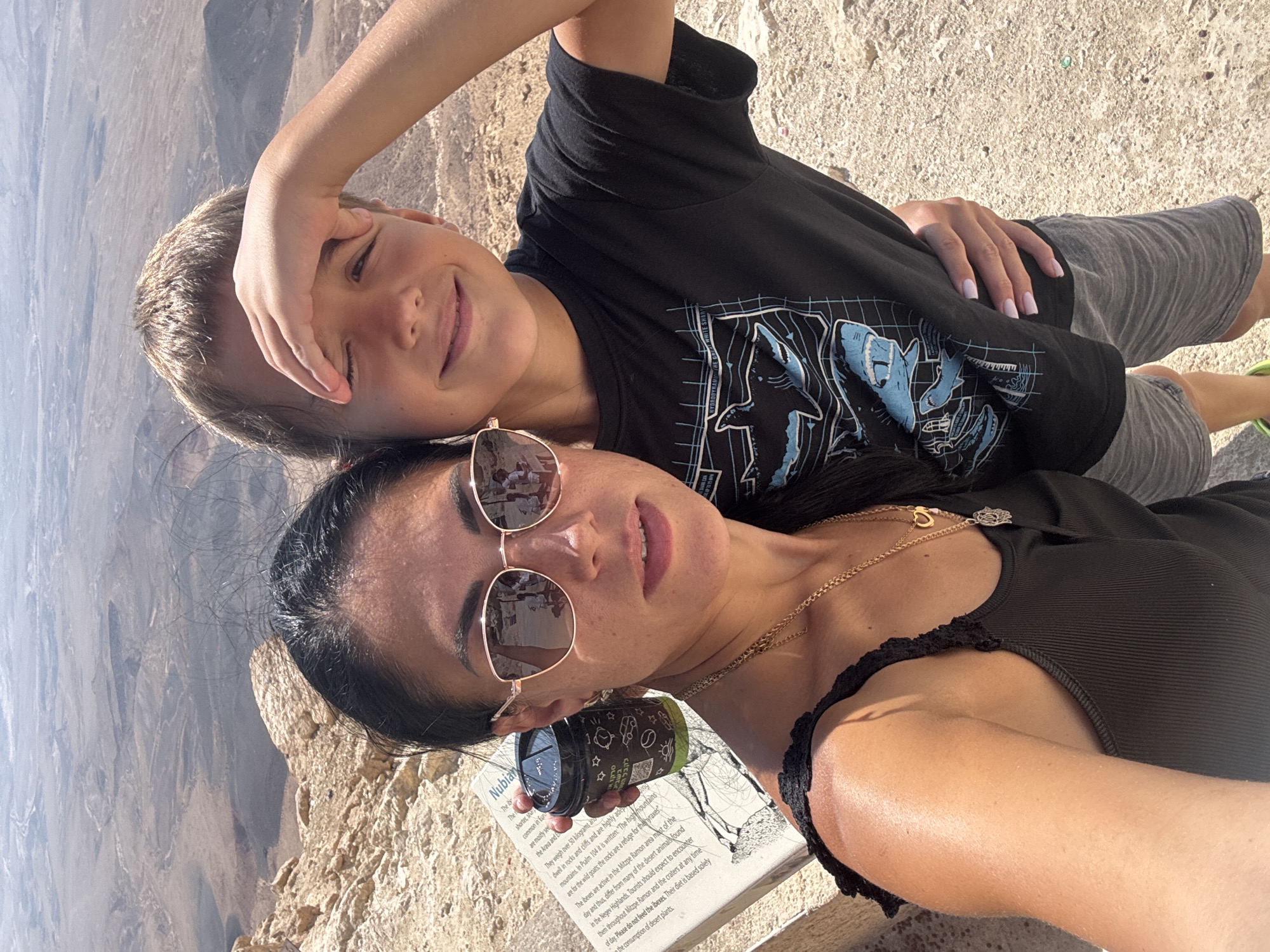 This is a selfie taken outdoors in Mitzpe Ramon, Israel. A woman with long dark hair and sunglasses is on the left, and a young boy with blond hair is on the right, leaning against her. The woman is wearing a black tank top and a gold necklace. She is holding a disposable coffee cup. The boy is wearing a black t-shirt with a shark graphic and grey shorts. He has his hand on his forehead, shielding his eyes from the sun. In the background, a vast desert landscape with mesas and canyons stretches out under a bright, hazy sky. A sign with text in English and Hebrew is partially visible on the left, providing information about the area. The ground is sandy and rocky.
