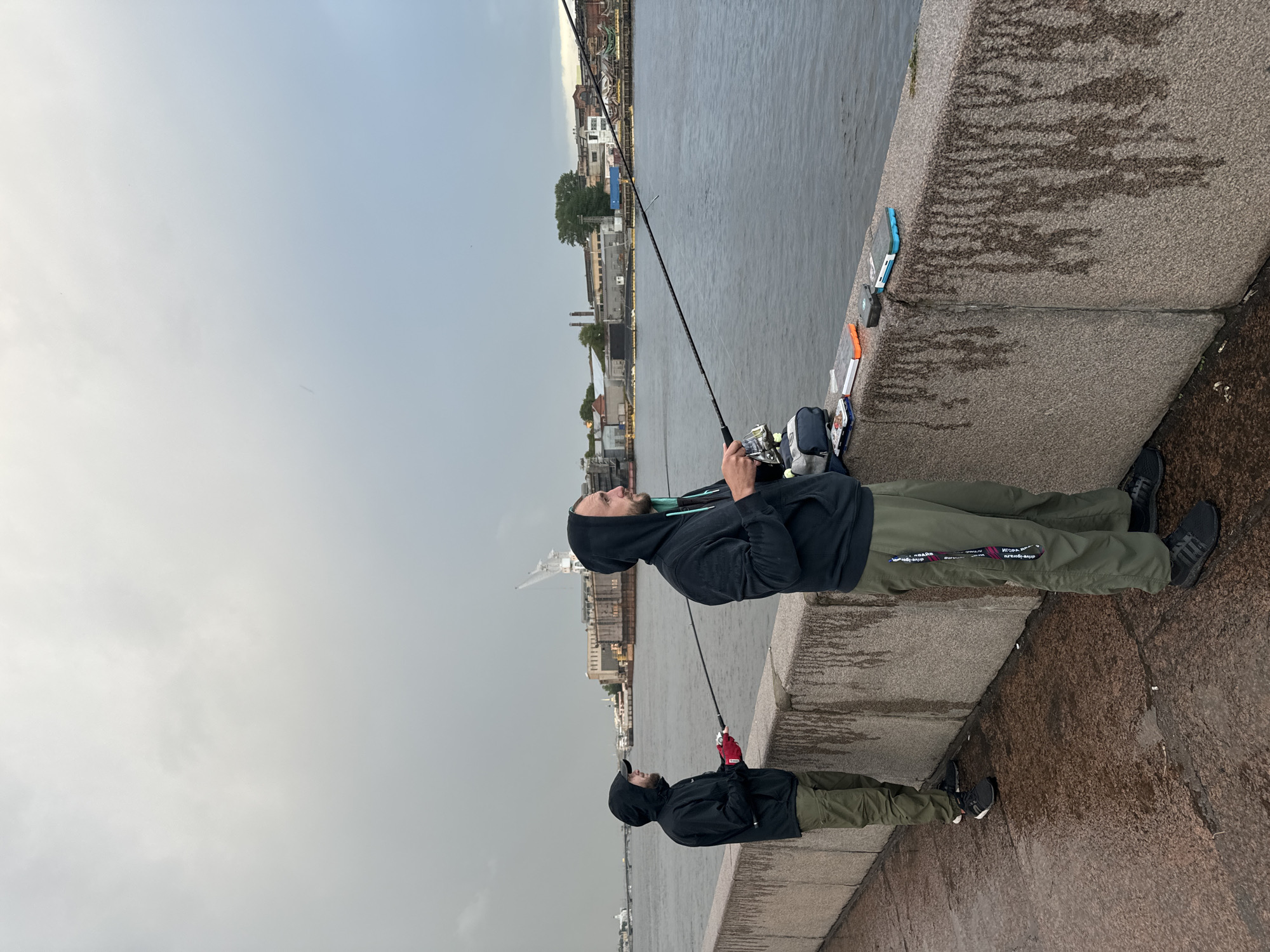 Two men are fishing on a granite embankment along the Neva River in Saint Petersburg, Russia. The sky is overcast and the water appears choppy. In the background, there are industrial buildings and a crane. The man on the left is wearing a black hoodie and green pants, holding a fishing rod with red gloves on. The man on the right is also wearing a black hoodie and green pants, holding a fishing rod. Several fishing tackle boxes are placed on the embankment. The ground is wet, indicating recent rain.