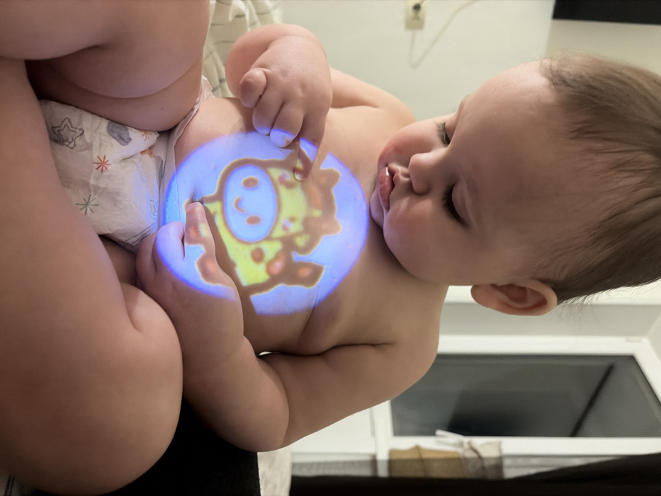 The image captures a close-up, angled view of a baby interacting with a projected image on their stomach. The baby, likely an infant or young toddler, is positioned on their back or held in such a way that their upper torso and head are visible, angled towards the right side of the frame. They are wearing a white disposable diaper adorned with star and possibly other subtle patterns.
A luminous, circular projection, predominantly blue around the edges, is cast onto the baby's bare abdomen. Within this glowing circle, a cartoon-like image of a yellow or light-orange animal, resembling a giraffe or a similar spotted creature, is visible. The animal has large, friendly-looking eyes and a wide, smiling mouth. The baby's left hand (from the viewer's perspective) is raised, with fingers gently extended towards the projected image, suggesting curiosity and engagement. One finger appears to be lightly touching the center of the projection.
The baby's head is turned slightly to the right, and their eyes are mostly closed or looking downwards, possibly in a state of calm concentration or drowsiness. Their mouth is slightly open, and their cheeks appear full and soft.
The background is softly out of focus, indicating an indoor setting. In the lower right corner, a white-framed object, possibly a window or a piece of furniture, can be discerned. The overall lighting in the room appears dim, which allows the projected image to stand out brightly on the baby's skin. No discernible text is visible in the image. The atmosphere conveyed is one of quiet play and gentle interaction.