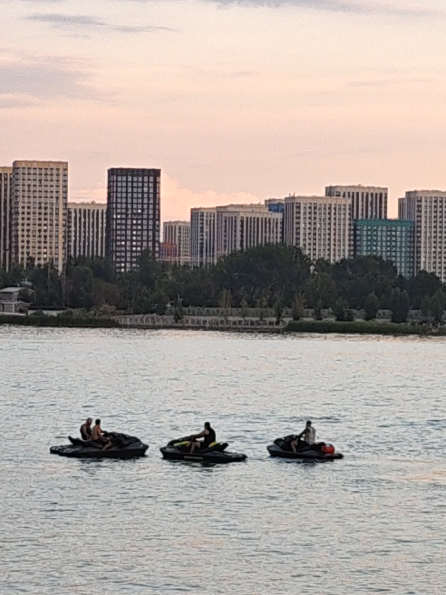 The image depicts a water scene with a cityscape in the background. In the foreground, three jet skis are visible on the water. The leftmost jet ski carries two individuals, one seated behind the other. The middle jet ski has one person seated and appears to be moving. The rightmost jet ski is occupied by a single person. The water is calm with ripples. In the middle ground, a shoreline is lined with trees. The background features a dense collection of tall buildings, characteristic of a city skyline. The sky above is a pale pink and grey, suggesting either dawn or dusk. The overall impression is one of recreational activity against an urban backdrop. This scene is likely located in Rostov-on-Don, Russia, where such waterfronts and modern architecture are common.