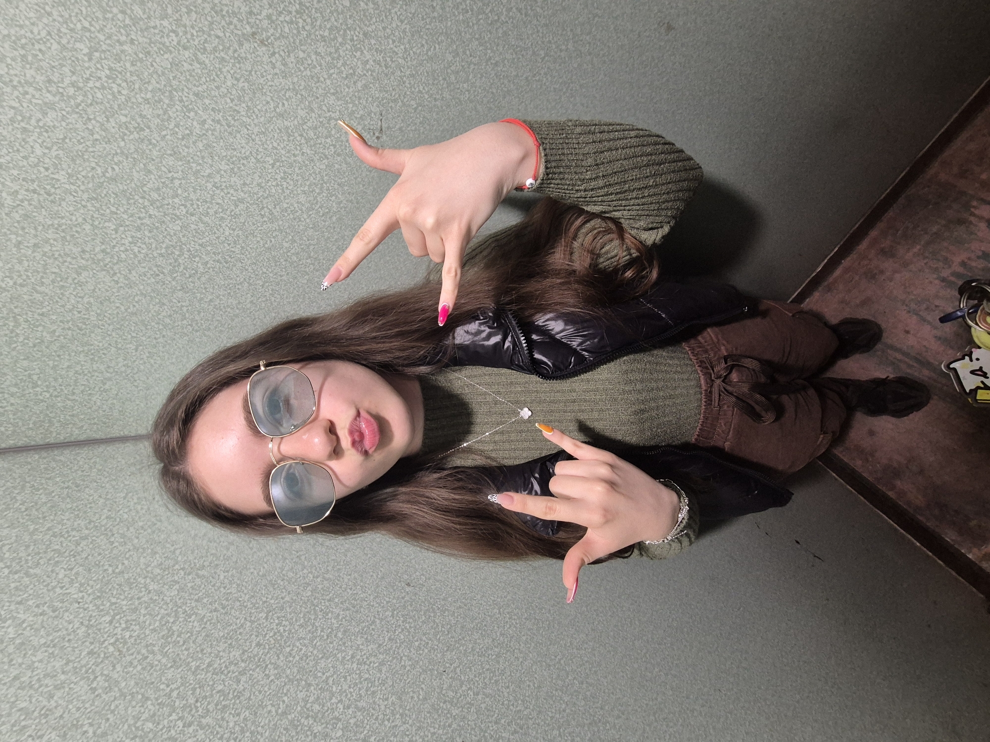 A young person, likely female, is captured in a diagonal, full-body shot, looking towards the camera with a slightly open mouth and a playful expression. She has long, dark brown hair and is wearing gold-framed sunglasses. Her attire includes a green knitted sweater, a black puffer vest, and brown pants.
Both of her hands are raised and forming a gesture where the thumb and pinky finger are extended, and the middle two fingers are curled down. Her long, well-manicured nails feature distinct designs and colors on each finger, such as gold, black and white patterns, hot pink, and orange. Accessories include a red string bracelet with a white bead on her left wrist, a silver-colored chain bracelet on her right wrist, and a silver-colored necklace with a white flower-shaped pendant.
The background is composed of two textured surfaces: a large, light green/grey stippled area covering most of the image, and a smaller, darker reddish-brown, wood-like surface in the lower right quadrant. A set of keys attached to a keyring with several small, colorful tags or charms is visible on the reddish-brown surface. One yellow tag shows some small, illegible black text or symbols. The lighting suggests an indoor environment.