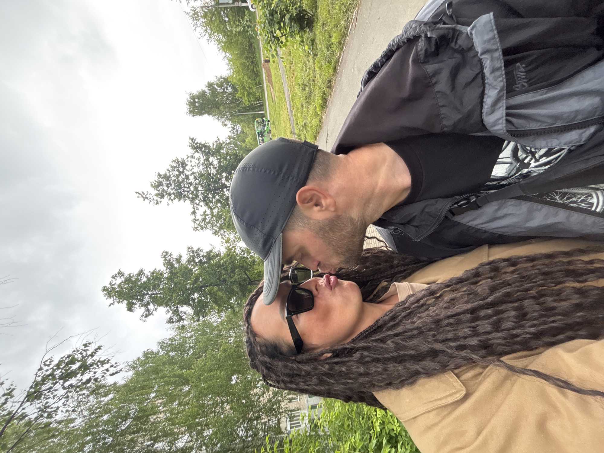 A man and a woman are captured in a close-up, outdoors, engaged in a kiss. The scene is located in the city of Chaikovsky, Russia. The woman, positioned on the left, has long, dark, textured hair styled in dreadlocks and wears dark, thick-framed sunglasses. She is dressed in a light brown or tan jacket over a lighter top, with her lips pursed for the kiss. The man, on the right, wears a dark baseball cap and a black and grey jacket over a black t-shirt, with stubble visible on his face. Their heads are tilted towards each other, and their lips are touching. The background features green foliage, including trees and bushes, indicative of a park-like or suburban setting. To the left, a light-colored building with windows is partially visible. To the right, a paved path or road extends, with a green bus and a partially visible light-colored car in the mid-ground. The sky is overcast with light grey clouds.