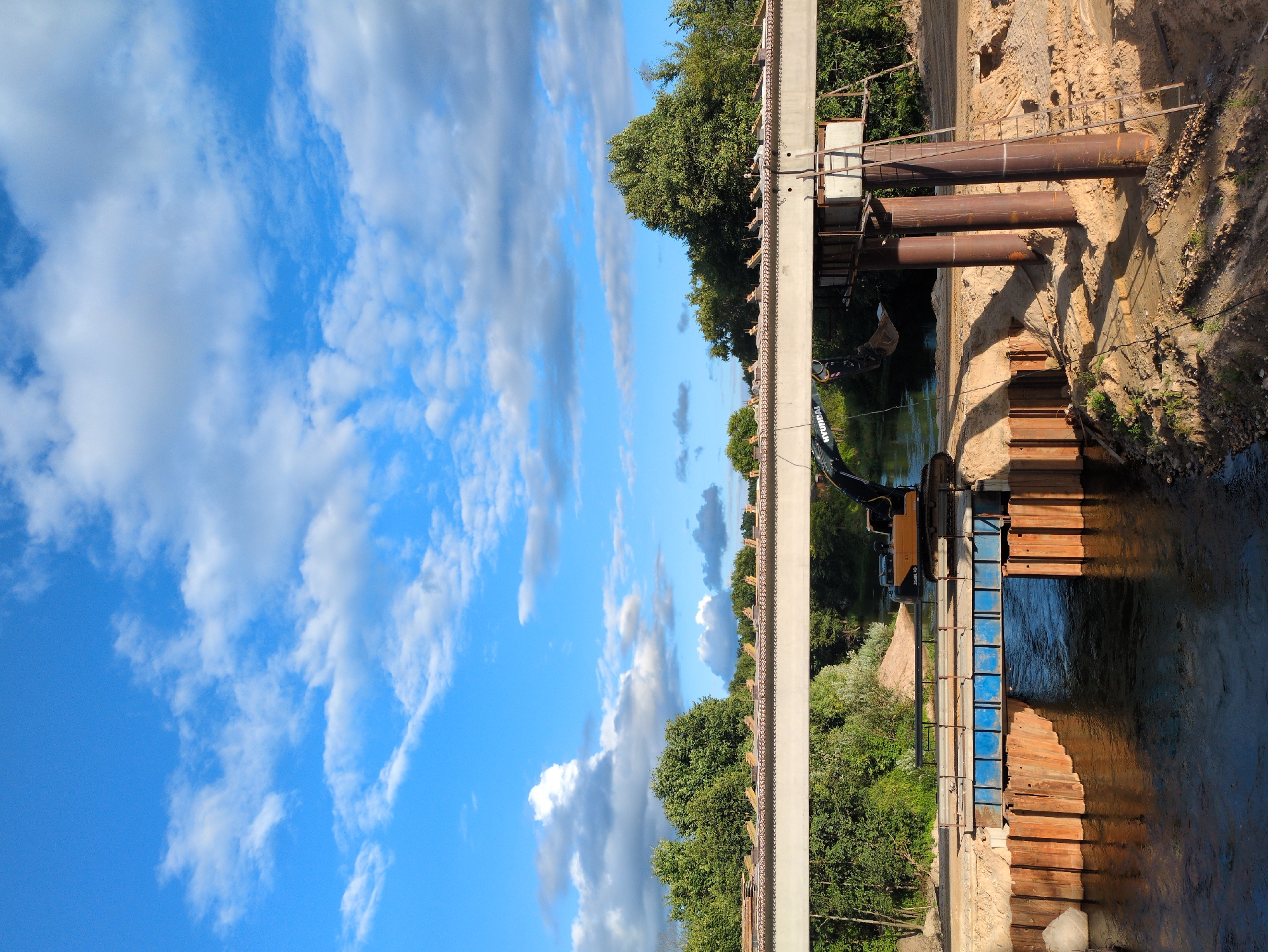 The image shows a construction site for a bridge over a river in Molochnaya Ferma, Russia. A yellow excavator is positioned on the riverbank, with its arm extended over the water. Construction materials, including steel pipes and wooden planks, are visible around the bridge structure. The river flows beneath the partially constructed bridge, which is supported by large concrete pillars and temporary wooden supports. Lush green trees line the riverbanks, and the sky is a bright blue with scattered white clouds. The scene appears to be a typical construction project in a rural or semi-rural setting, with the focus on the engineering and infrastructure development.
