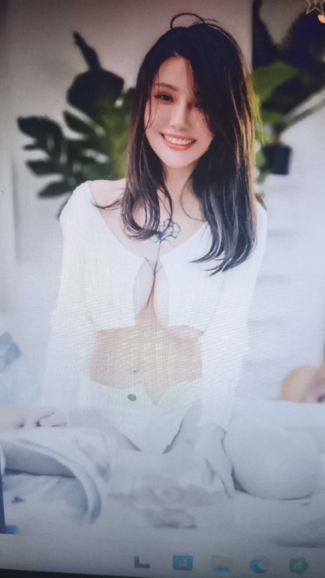 The image shows a woman with dark hair smiling at the camera. She is wearing a white top and white bottoms. A tattoo is visible on her chest. Behind her, there is a large green plant. The image appears to be taken from a computer screen displaying the picture. The location is not discernable from the image and can not be placed in Nowy Tomyśl, Poland.