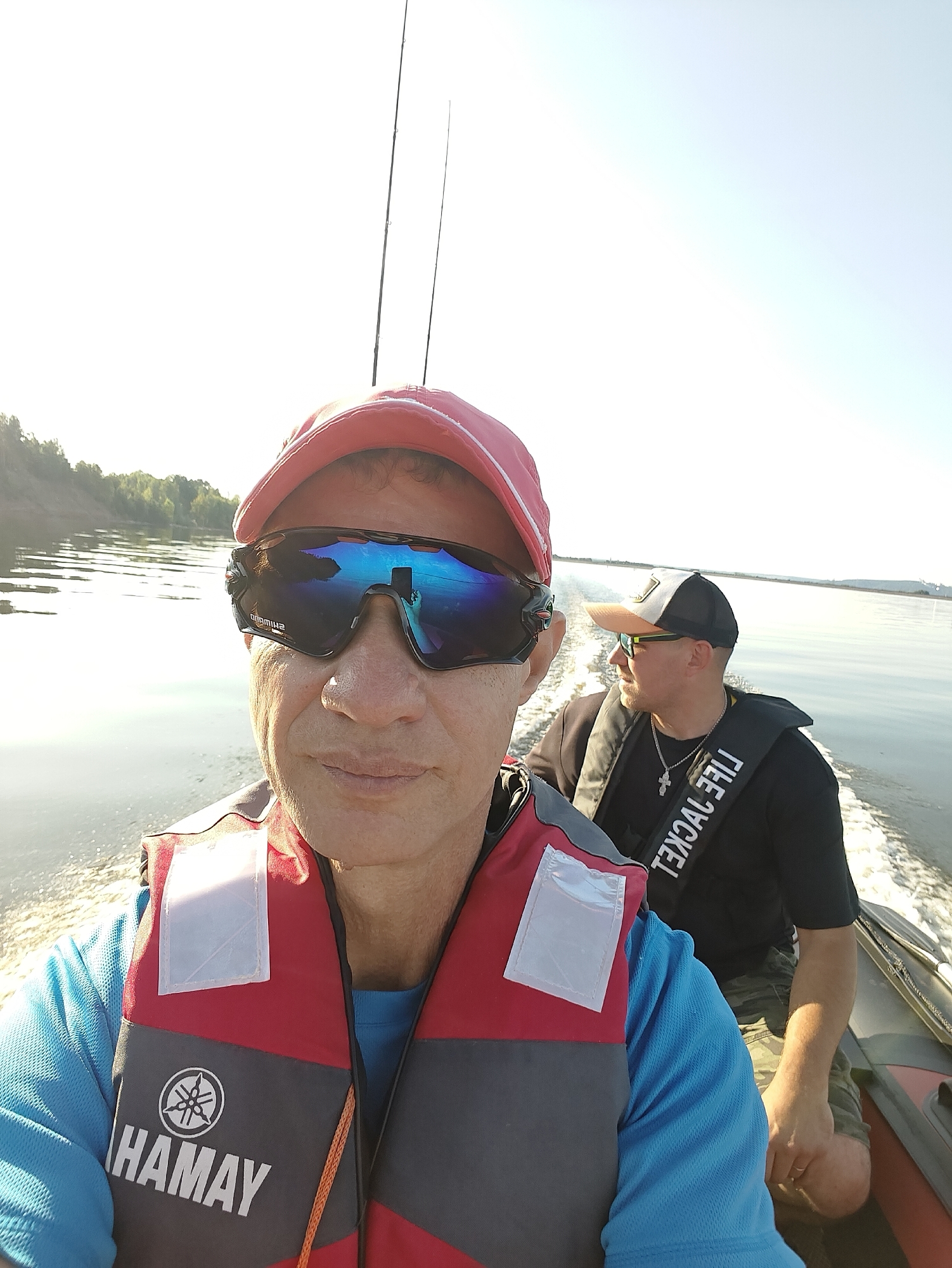 In the foreground, a person wearing a red and gray life vest with a Yamaha logo and a blue shirt is taking a selfie on a boat. They are also wearing a red baseball cap and reflective sunglasses. Behind them, another person in a black shirt, life vest, and a baseball cap is visible, looking to the side. Both individuals appear to be on a boat moving through calm water under a clear sky, with trees and a riverbank visible in the distance on the left. The scene takes place on the river in Noviy, Russia.