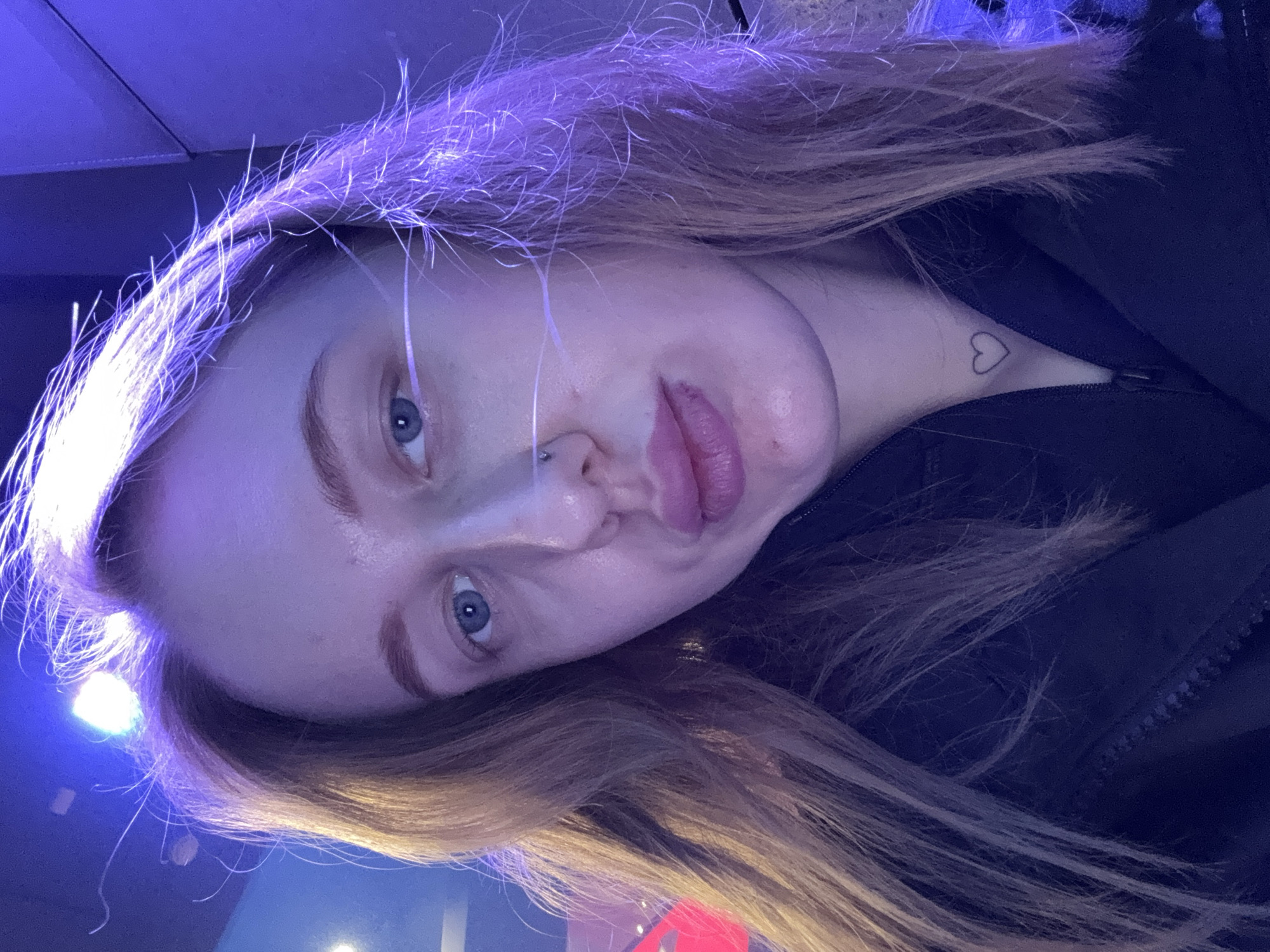 The image shows a fair-skinned woman with blue eyes and light brown hair. She has a nose piercing on the left side. A small heart tattoo is visible on her neck. She is wearing a dark-colored jacket. The lighting is predominantly blue and purple. The location is not identifiable as being in Ol'ginskaya, Russia.