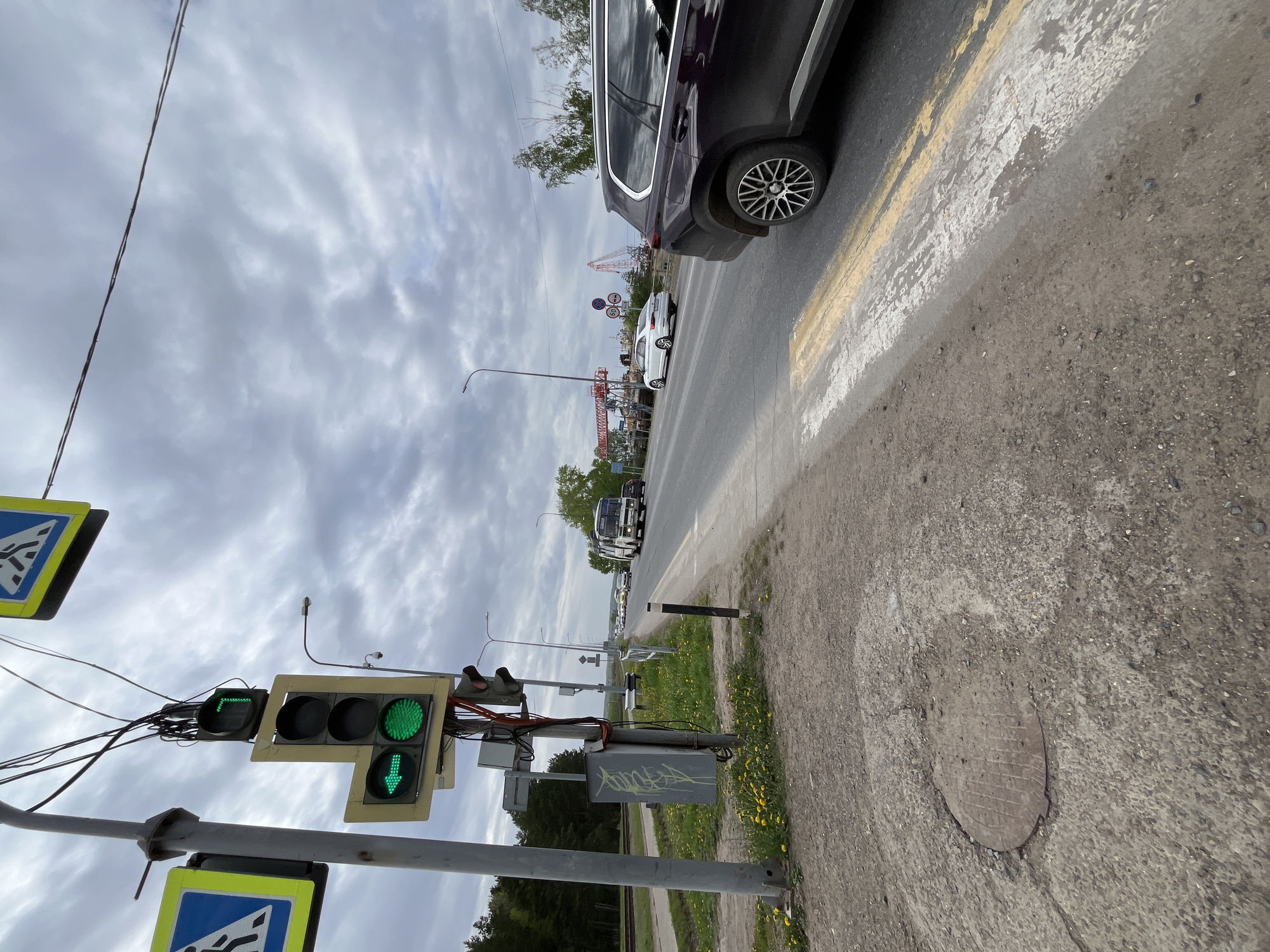 The image depicts a street scene in Chaikovsky, Russia, taken from a low vantage point near a sidewalk. A traffic light displaying a green light for both straight and left turns is visible on a pole with pedestrian crossing signs. Several cars are lined up on the road, including a dark SUV and a white sedan. A ferris wheel and other amusement park structures are visible in the distance. The sky is overcast with grey clouds. The sidewalk is made of concrete and shows signs of wear. There are yellow flowers growing along the edge of the sidewalk.