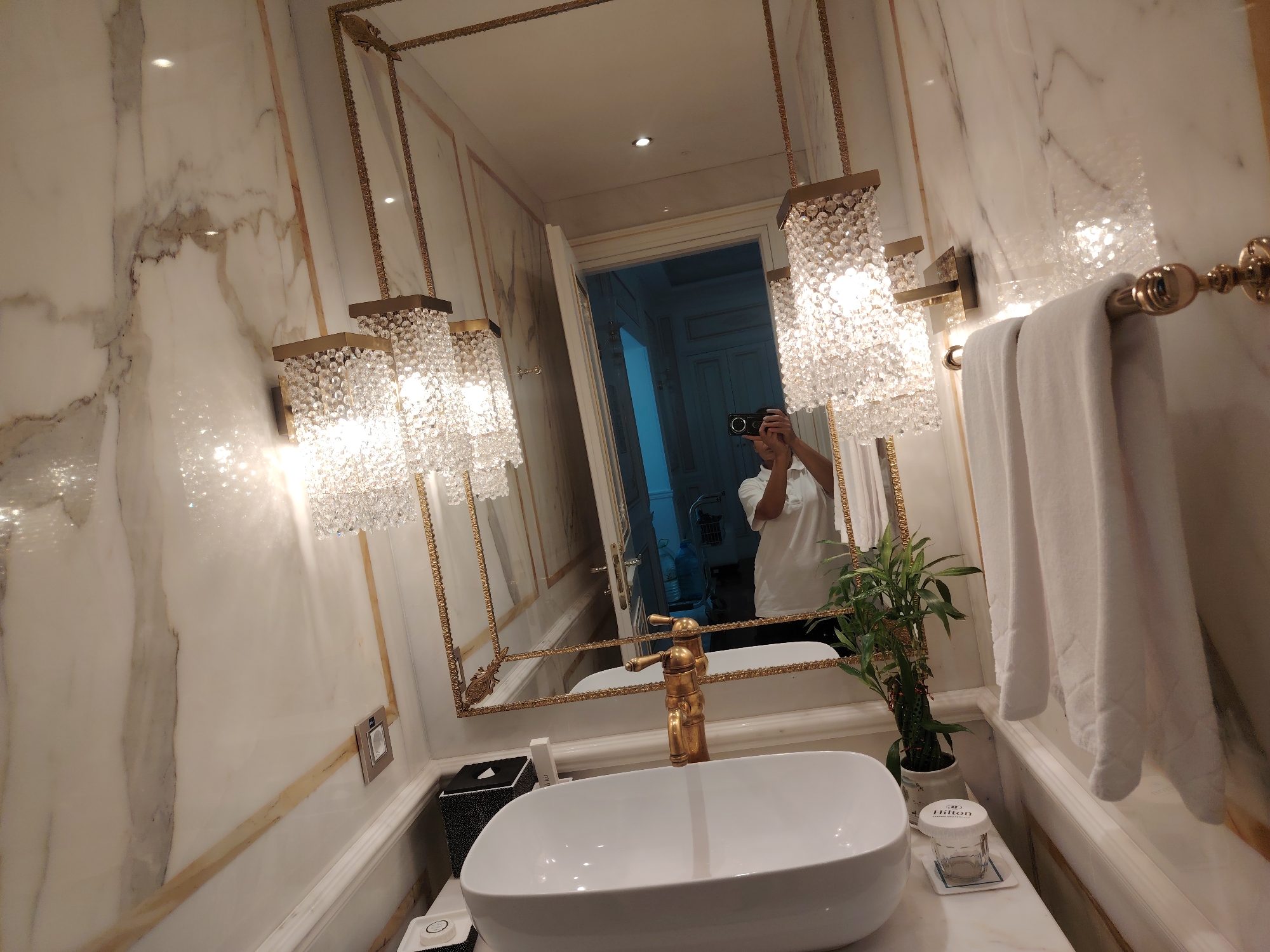A first-person perspective shot captures a luxurious bathroom in Konigil, Uzbekistan. The walls are clad in white marble with prominent golden veins and trim, reflecting the light from ornate crystal chandeliers hanging from the ceiling. A large, gold-framed mirror dominates the scene, reflecting a person holding a camera, presumably the photographer. The person is wearing a white shirt.
In the foreground, a white porcelain sink with a vintage-style gold faucet sits on a marble countertop. To the left of the sink is a black tissue box with a white pattern. To the right, a small potted bamboo plant adds a touch of green. A white folded towel hangs on a gold rack on the right wall. The overall impression is one of opulence and attention to detail.