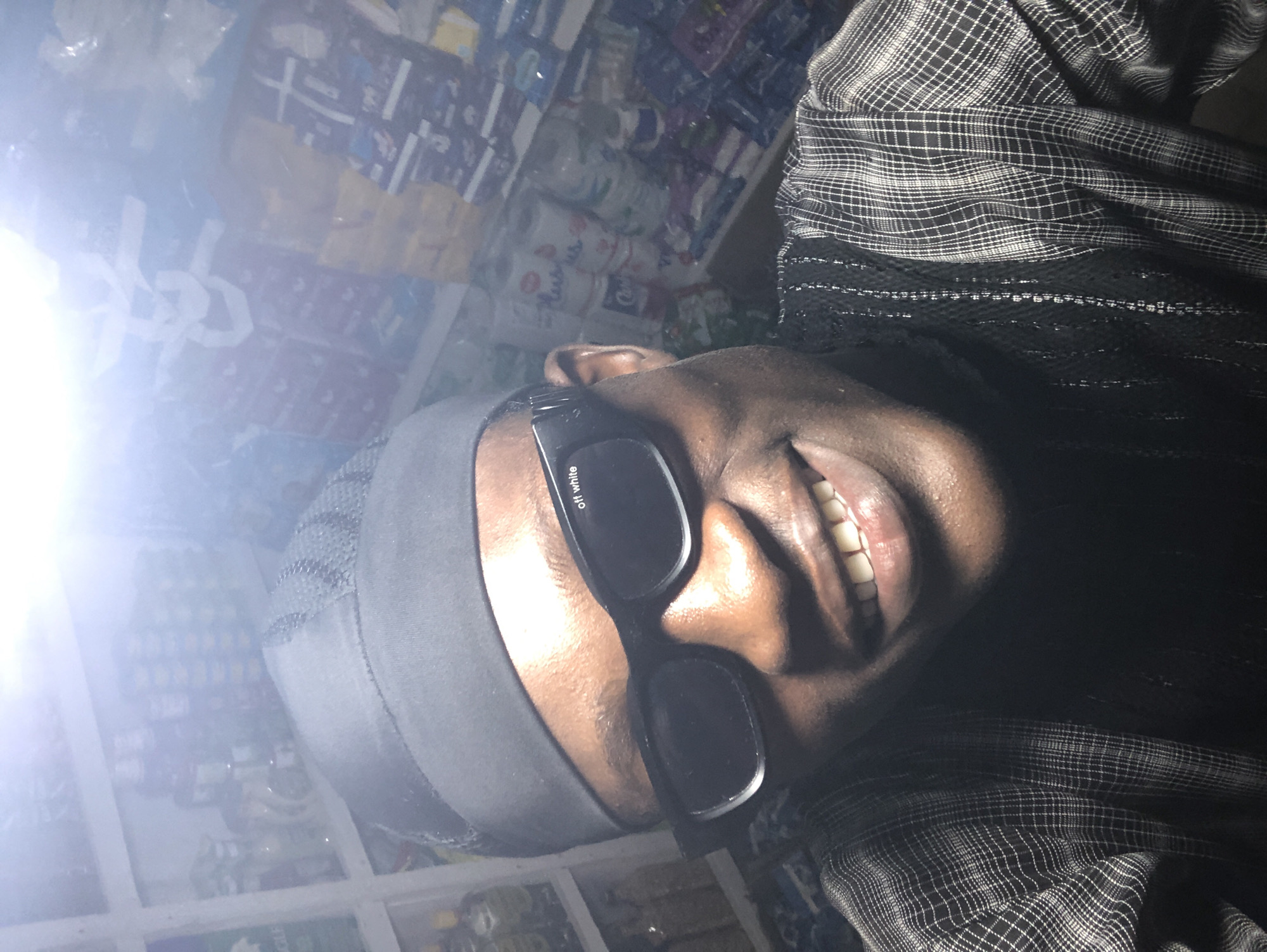 In Ado, Nigeria, a close-up, selfie-style photograph captures a young Black man wearing a black durag and black-framed sunglasses with "off white" written on them. He is smiling, revealing his teeth. He is also wearing a black and white checkered shirt over a black garment. The background appears to be a store, with shelves filled with various products, including bottles and packaged goods, suggesting a retail environment. The lighting in the image is bright and somewhat overexposed, particularly from a light source directly above and to the left.