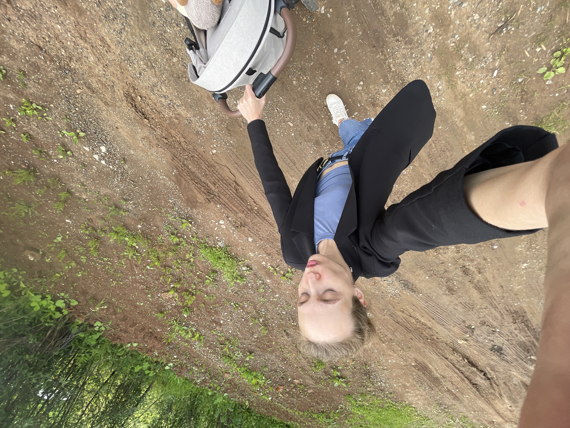 A person is standing on a dirt path next to a baby stroller. The person is wearing a black blazer over a blue tank top and blue jeans, with white sneakers. They are holding the handle of the stroller with their right hand, and their left arm is extended towards the camera, holding a cell phone which is taking the picture. The person's eyes are closed, and their mouth is slightly open, with a gentle expression on their face. A teddy bear is visible in the stroller. The path is made of dirt and gravel, with some tire tracks visible. There are patches of green grass and plants growing along the sides of the path, and trees in the background. The lighting suggests it is daytime, and the overall atmosphere is casual and relaxed. The location is Sharkan, Russia. No text is visible in the image.