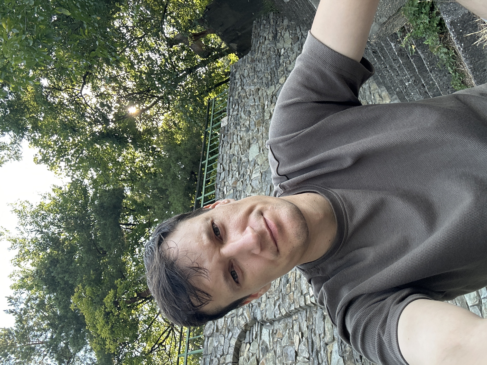 A man is taking a selfie outdoors in Makiv, Ukraine. He is positioned in the lower center of the frame, looking directly at the camera with a neutral expression. He has short, dark hair and is wearing a dark gray t-shirt. Behind him, a stone wall forms a textured backdrop. To the left of the wall, there are visible stone steps, and to the right, overgrown greenery and more stone steps are present. Above the wall, the branches and leaves of mature trees dominate the upper portion of the image, with sunlight filtering through the canopy. The sky is visible in the upper right corner.