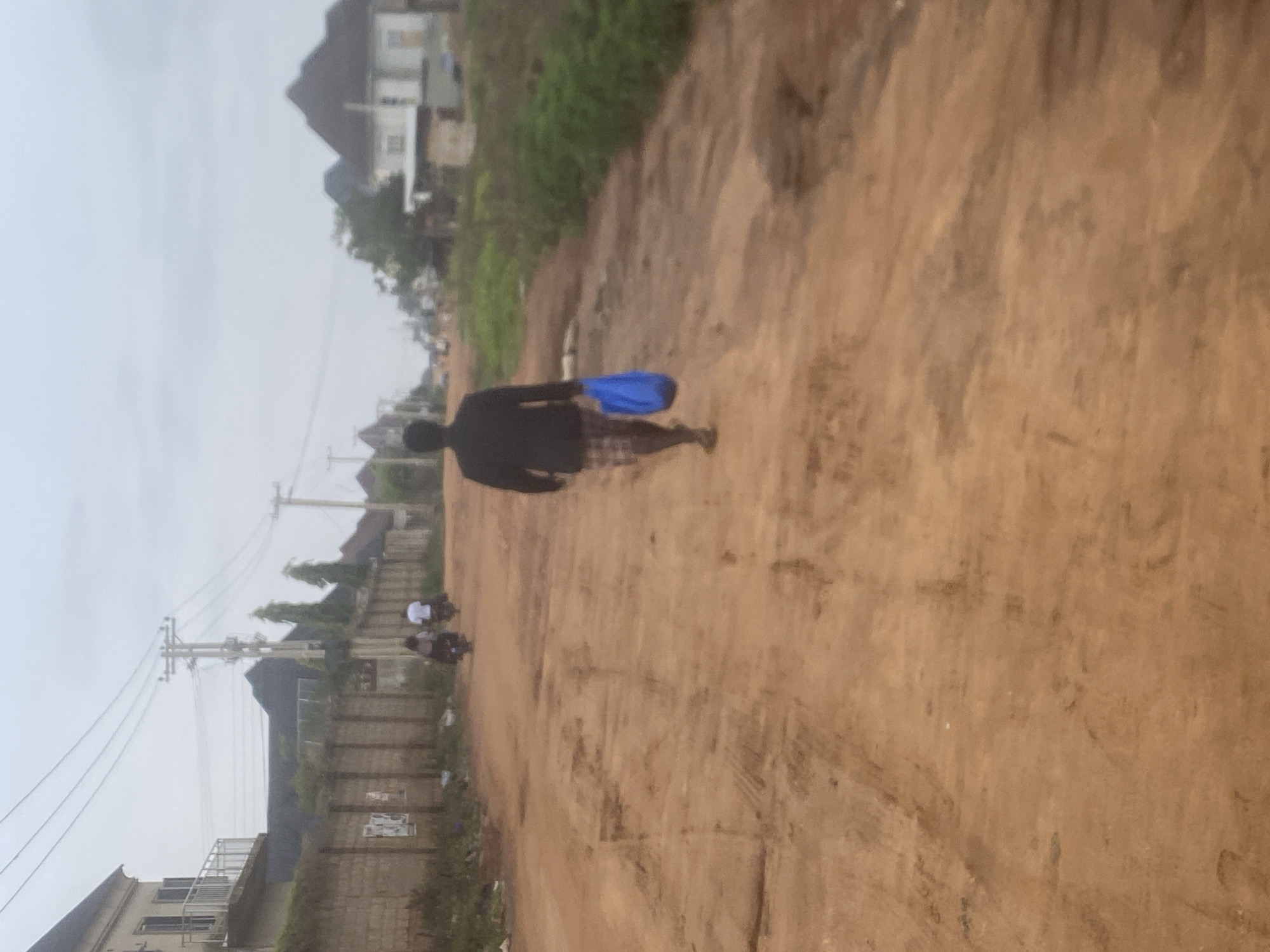 A person wearing a dark jacket and checkered shorts walks away from the camera on a dirt road in Iddo Tudunwada, Nigeria. The person is carrying a blue bag in their right hand. The road is unpaved and appears to be in a rural or developing area, with some puddles and uneven terrain. In the background, several houses with pitched roofs are visible, as well as utility poles and power lines. Two people are seen riding motorcycles further down the road. The sky is overcast.