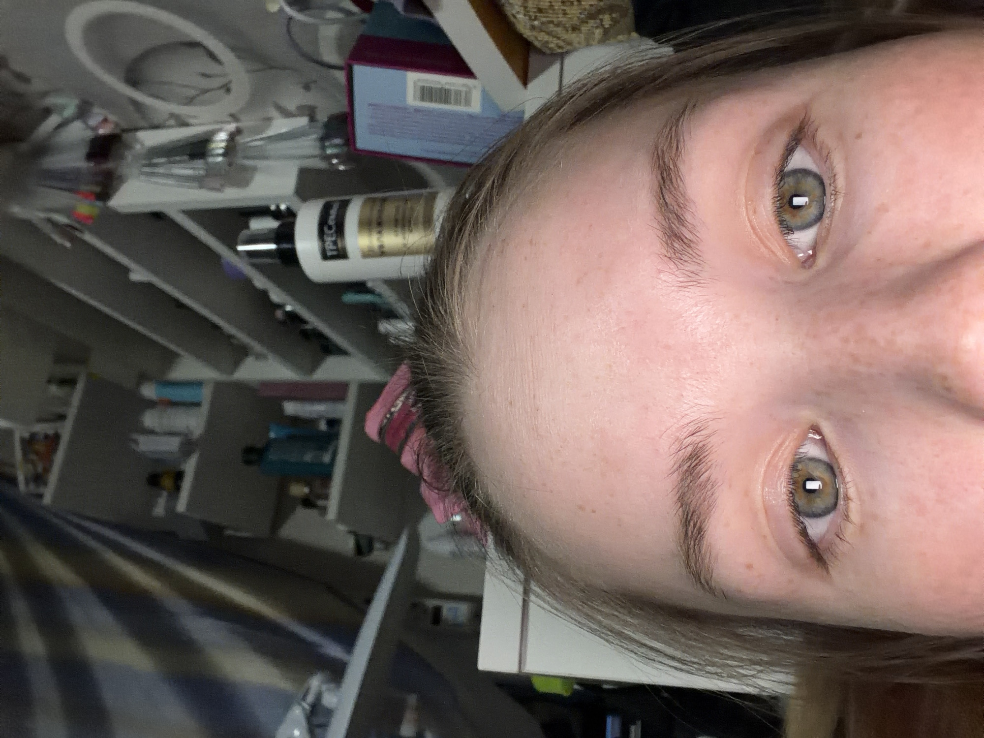 The image is a close-up shot of a person's face, primarily focusing on the eyes, eyebrows, and forehead. The eyes appear to be a hazel or gray-green color. The background consists of a cluttered shelf containing various bottles and containers, likely personal care products. There is also a visible light fixture and a striped pattern suggestive of bedding. The location is not possible to determine from the image.