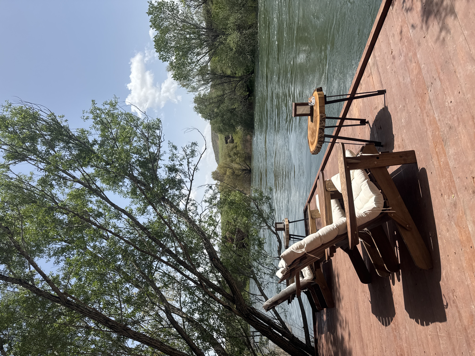 On a sunny day in Avanos İlçe Merkezi, Turkey, a serene waterfront scene unfolds. The image captures a wooden deck extending over a river. Two wooden chairs with white cushions are positioned on the deck, offering a comfortable spot to relax. A small, round table made from a tree slice stands nearby. The river flows alongside the deck, reflecting the surrounding greenery and sky. Trees line the opposite bank, adding to the natural ambiance. The sky is blue with scattered clouds.