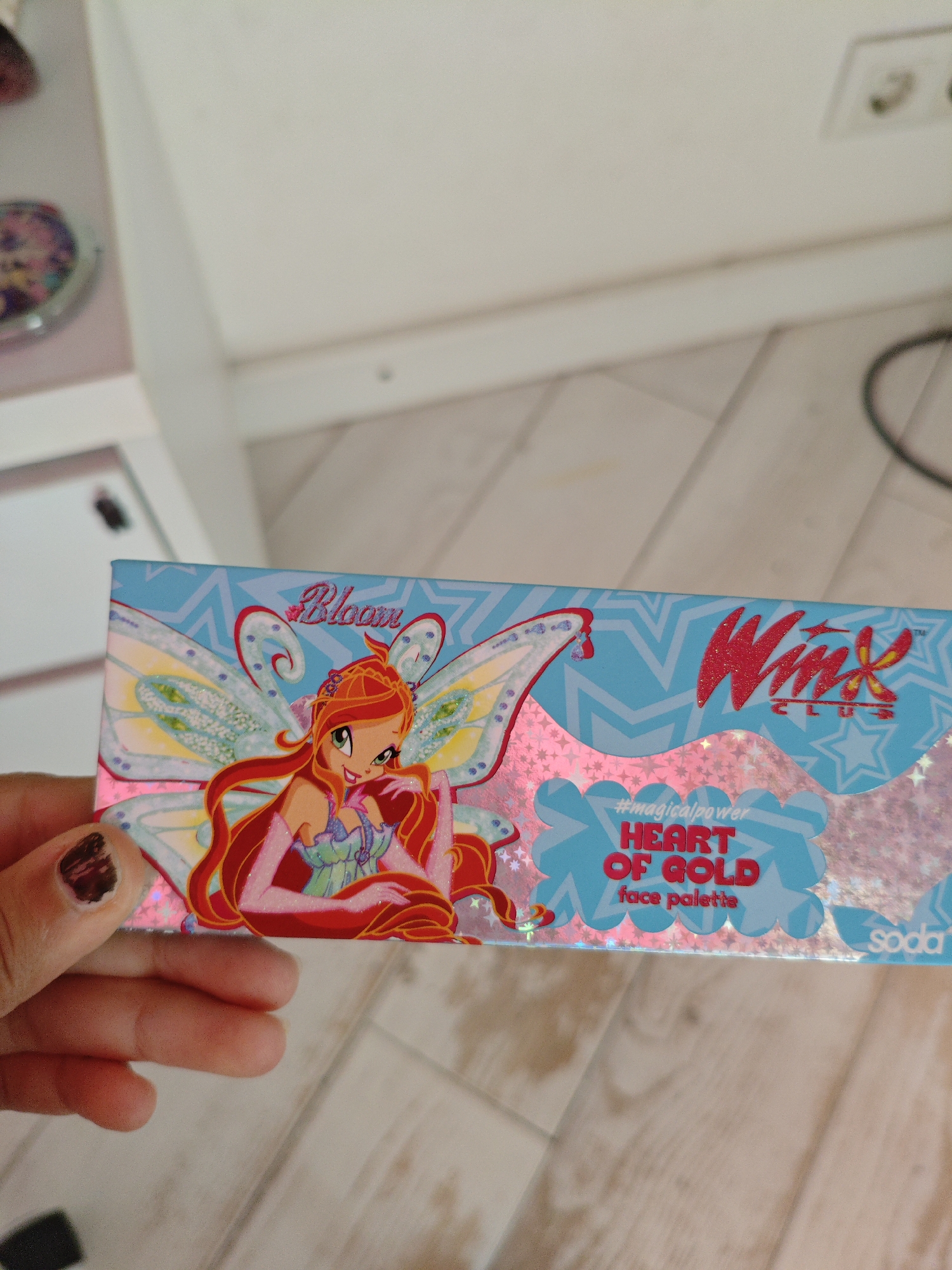 An overhead shot shows a hand holding a makeup palette box. The box features cartoon characters with wings and the words "Bloom Club," "#magicalpower," "HEART OF GOLD," and "face palette." The background appears to be a wooden floor. The location is identified as Трудовское сельское поселение, Ukraine.