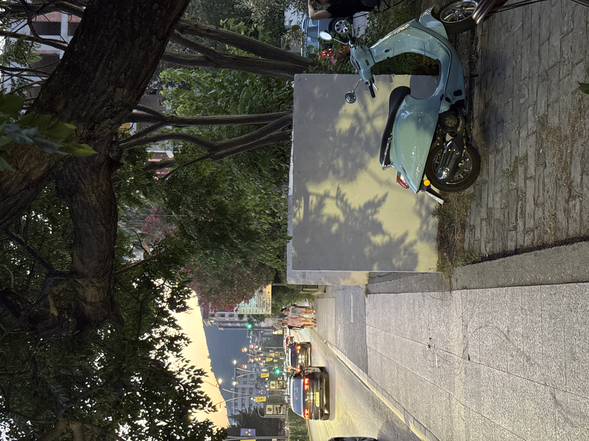 The image depicts a street scene in Budva, Montenegro, near the Old Town. A pale blue scooter is parked on a brick-paved area to the right of a sidewalk. Behind the scooter, a light-colored wall casts shadows. The sidewalk, paved with grey stone tiles, runs along a street with several cars driving on it. The street is lined with buildings and trees. In the background, streetlights are visible, and hills are silhouetted against an orangey sky, suggesting it is either sunrise or sunset. A group of people are walking on the sidewalk in the middle ground. The overall impression is of a warm, late afternoon or early evening urban setting.