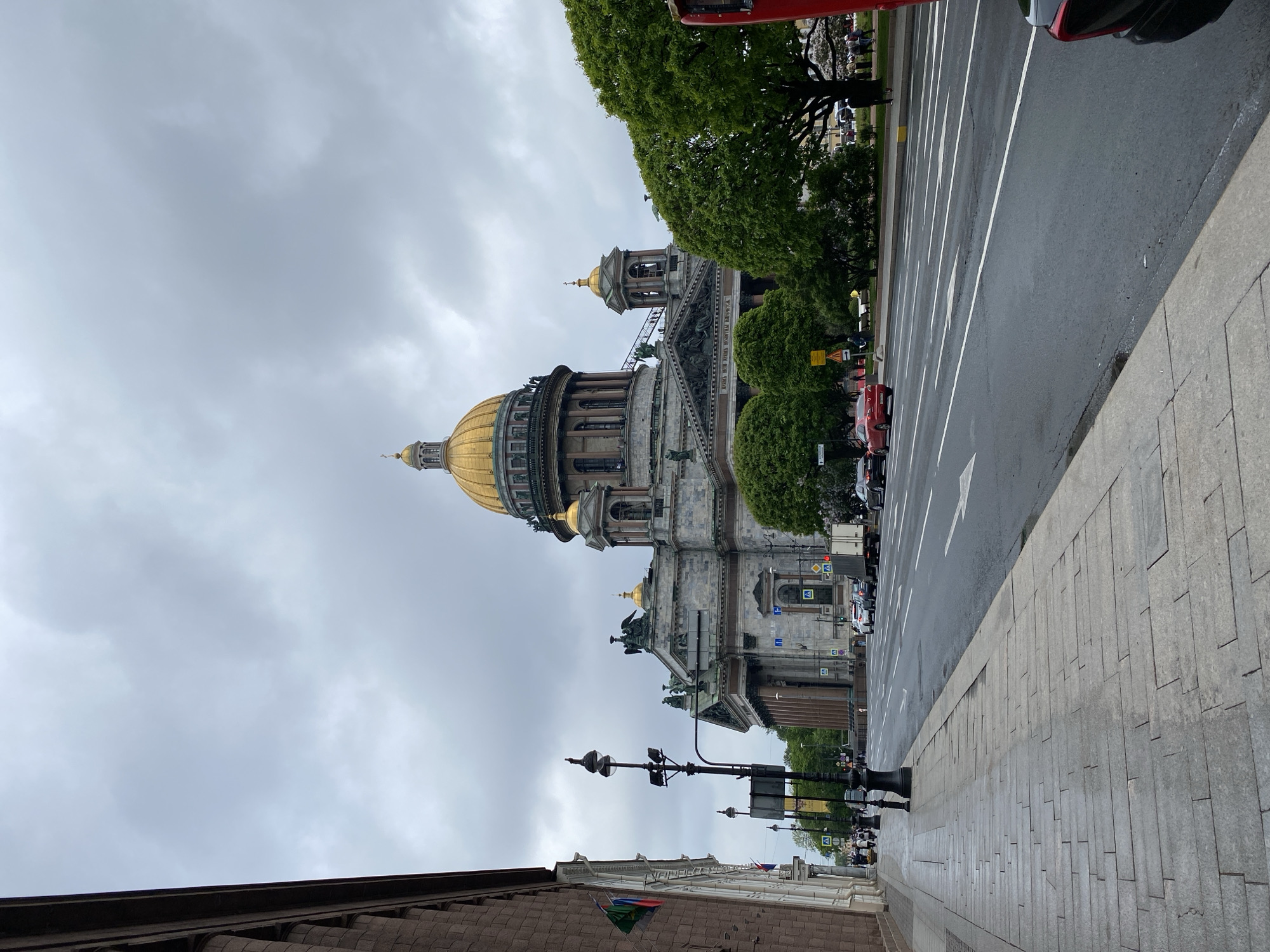 The image depicts Saint Isaac's Cathedral in Saint Petersburg, Russia. The cathedral is viewed from street level, showcasing its golden dome and facade. The sky is overcast. To the right is a street with marked lanes and a sidewalk with a lamppost. There is also a sidewalk on the left.