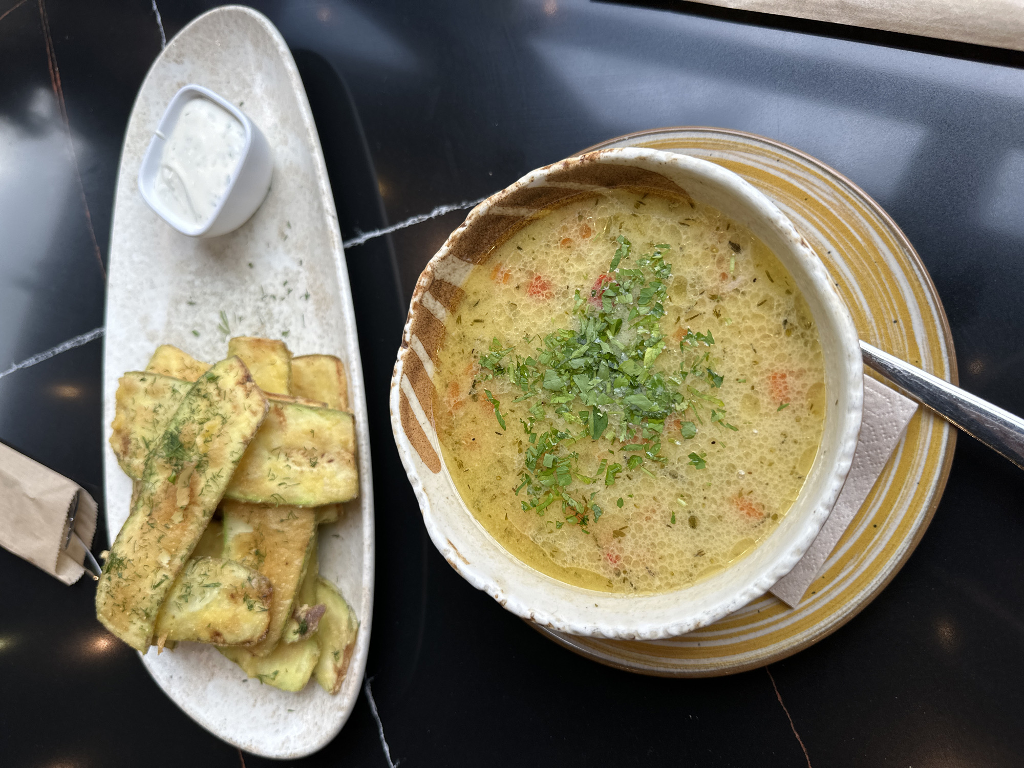 The image shows a table with a bowl of soup and a plate of fried zucchini with a dipping sauce. The soup is a light yellow color and has green herbs sprinkled on top. The zucchini is golden brown and also has green herbs sprinkled on top. The dipping sauce is white. The table is black with a marble pattern. The location is not specified as being in Montana, Bulgaria.