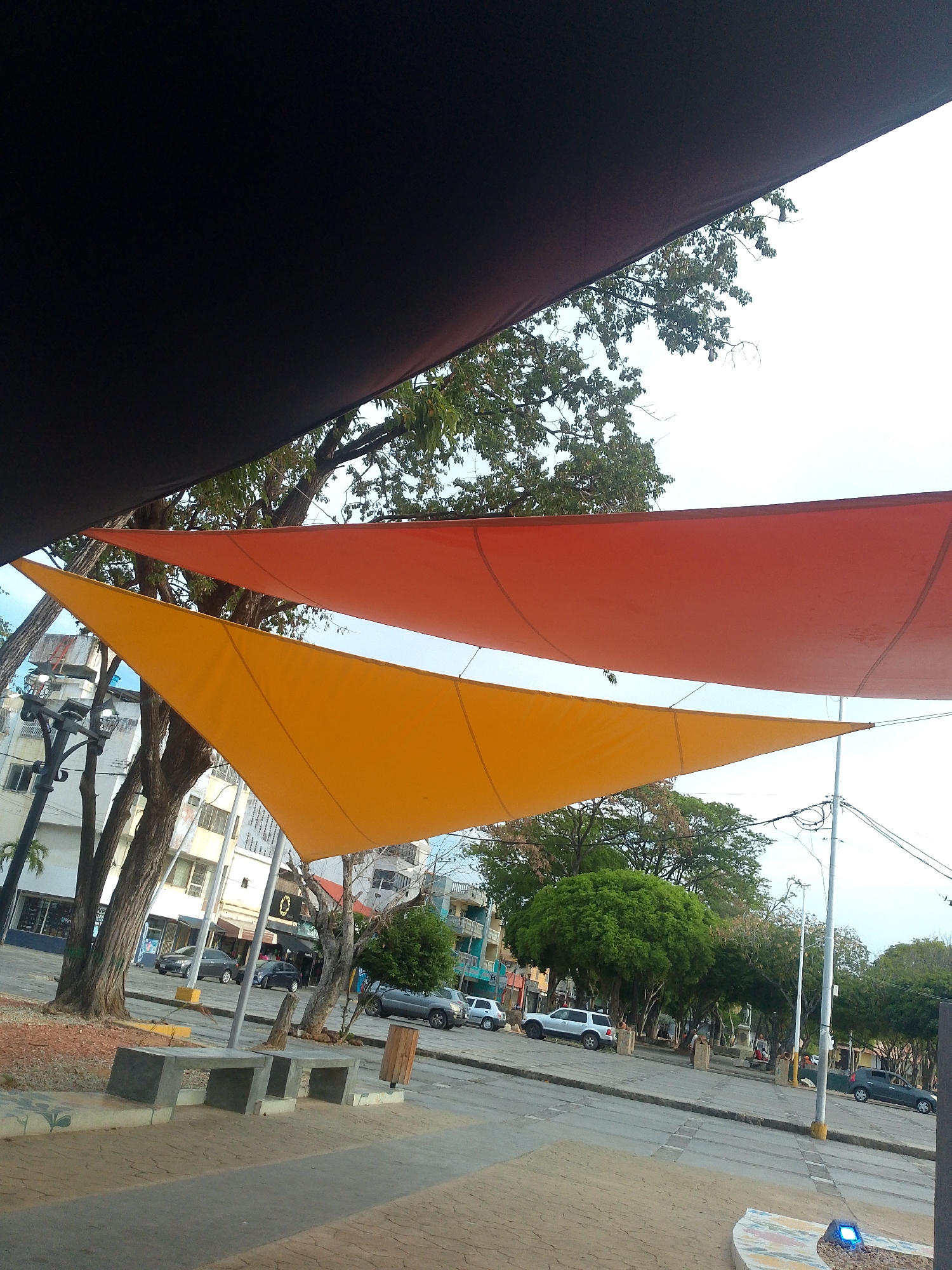 The image depicts a public space in Juan Griego, Venezuela, with triangular sunshades providing cover. One shade is orange, and the other is yellow. A third, black shade is partially visible at the top left. The scene includes trees, benches, a light post, and parked cars. Buildings are visible in the background. The location appears to be a town square or a similar pedestrian area. There are no people visible.