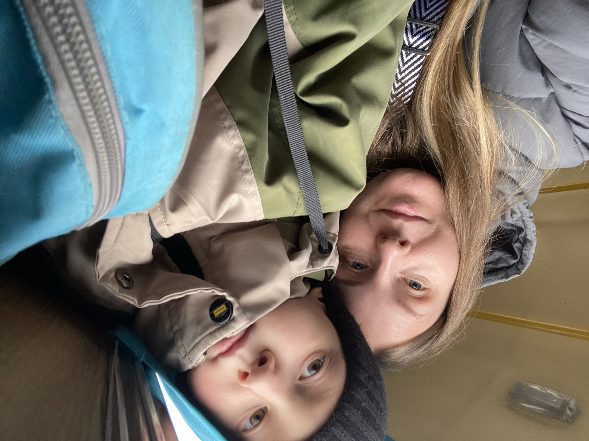 The image, taken from a low angle and rotated 90 degrees clockwise from its original orientation, captures an adult and a young child looking upwards towards the camera. The scene appears to be an enclosed indoor space, possibly within a vehicle like a train or bus, suggested by the warm, plain background and a blurry overhead light fixture seen in one of the cropped hints. This setting is located in Chaikovsky, Russia.
The adult, positioned slightly behind and above the child, has fair skin and light brown or blonde hair. She is wearing a grey hooded puffer jacket and a two-toned jacket, which is olive green on the upper part and khaki below. A strap with a black and white chevron pattern is visible near her shoulder.
The young child, closer to the viewer, has fair skin and dark eyes. They are wearing a dark grey or black knitted beanie and a khaki jacket. A notable detail on the child's jacket is a small, circular yellow and black patch that clearly displays the text "BOMBER". Both individuals maintain neutral expressions, looking directly into the lens.
To the far left of the frame, a bright blue object with a grey zipper is prominent, possibly part of a backpack or another garment. Another grey strap runs vertically in the center of the image. The attire of both individuals, featuring jackets and a beanie, suggests cool weather.