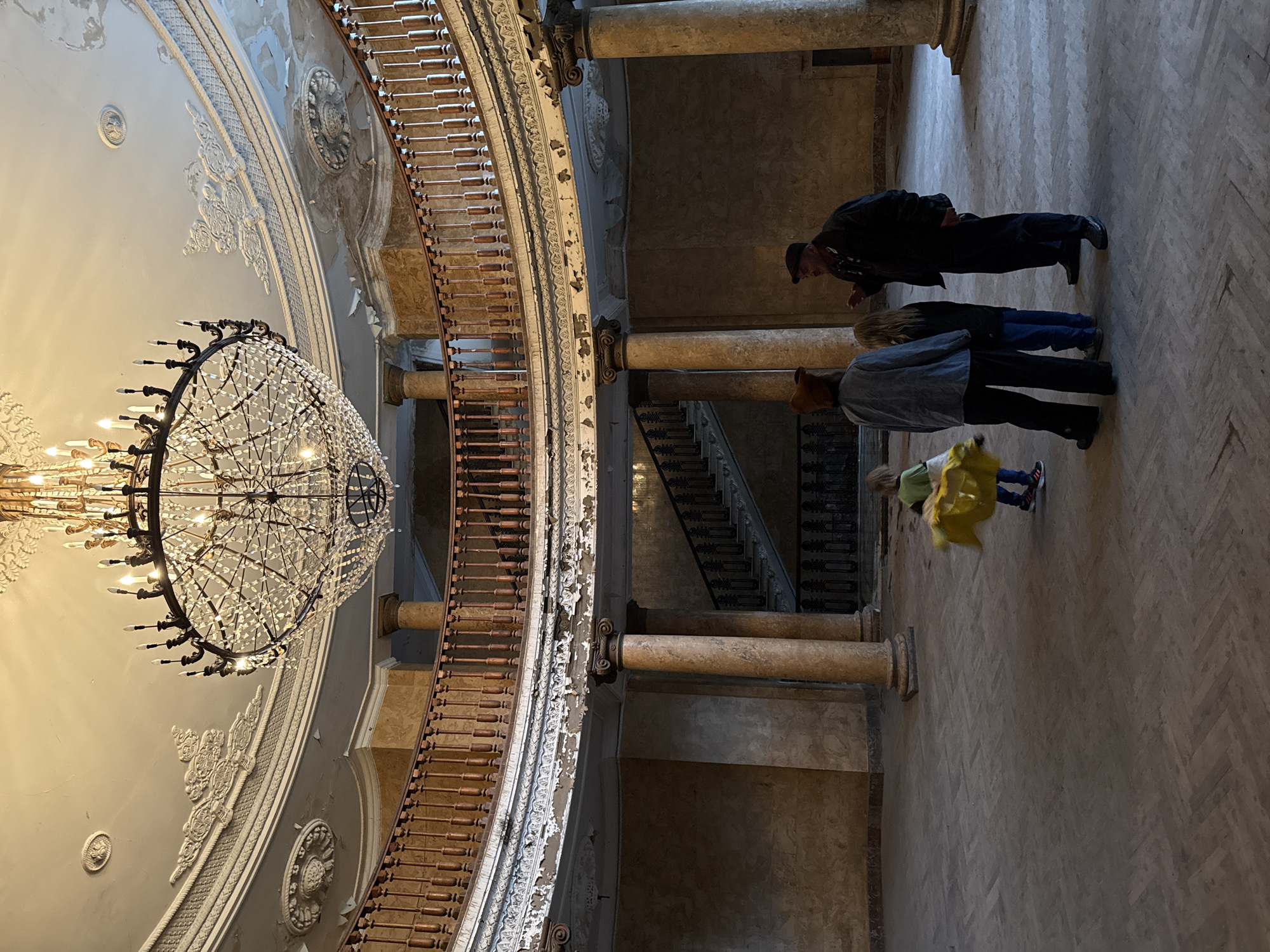 The image depicts the interior of an ornate building, likely a historical structure, in Tskaltubo, Georgia.
Several individuals are present, including an elderly man wearing a dark suit and a cap, who appears to be observing something closely. A group of three individuals – adults and a child – stand near the man. The child is wearing a yellow coat and blue pants. They stand near marble columns and another flight of stairs is visible in the distance.
The interior features a grand, multi-tiered balcony with intricate railings and decorative detailing. A large, ornate chandelier hangs from the ceiling, which is adorned with classical motifs and decorative plasterwork. The walls exhibit signs of aging and disrepair, with peeling paint and weathered surfaces. The floor has a herringbone pattern.