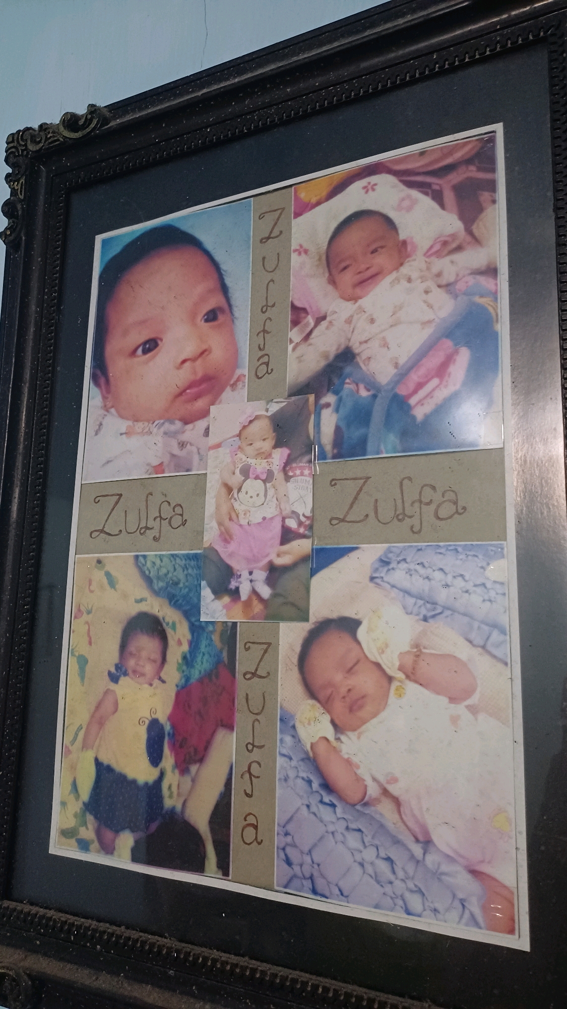 This is a framed photograph containing multiple smaller images of a baby. The frame is dark with ornate corner details. The photographs are arranged in a grid-like pattern.
In the top left, a close-up of a baby's face is visible. To its right, another baby is smiling, lying on its back. A smaller photograph in the center shows a baby wearing a pink dress with a Mickey Mouse motif. Below the first two images, the word "Zulfa" is written. To the right of the central image, another "Zulfa" is written. In the bottom left, a baby is wearing a yellow and blue outfit. To its right, a baby is sleeping on its back.
The photographs appear to be from the city of Kowangan, Indonesia.
