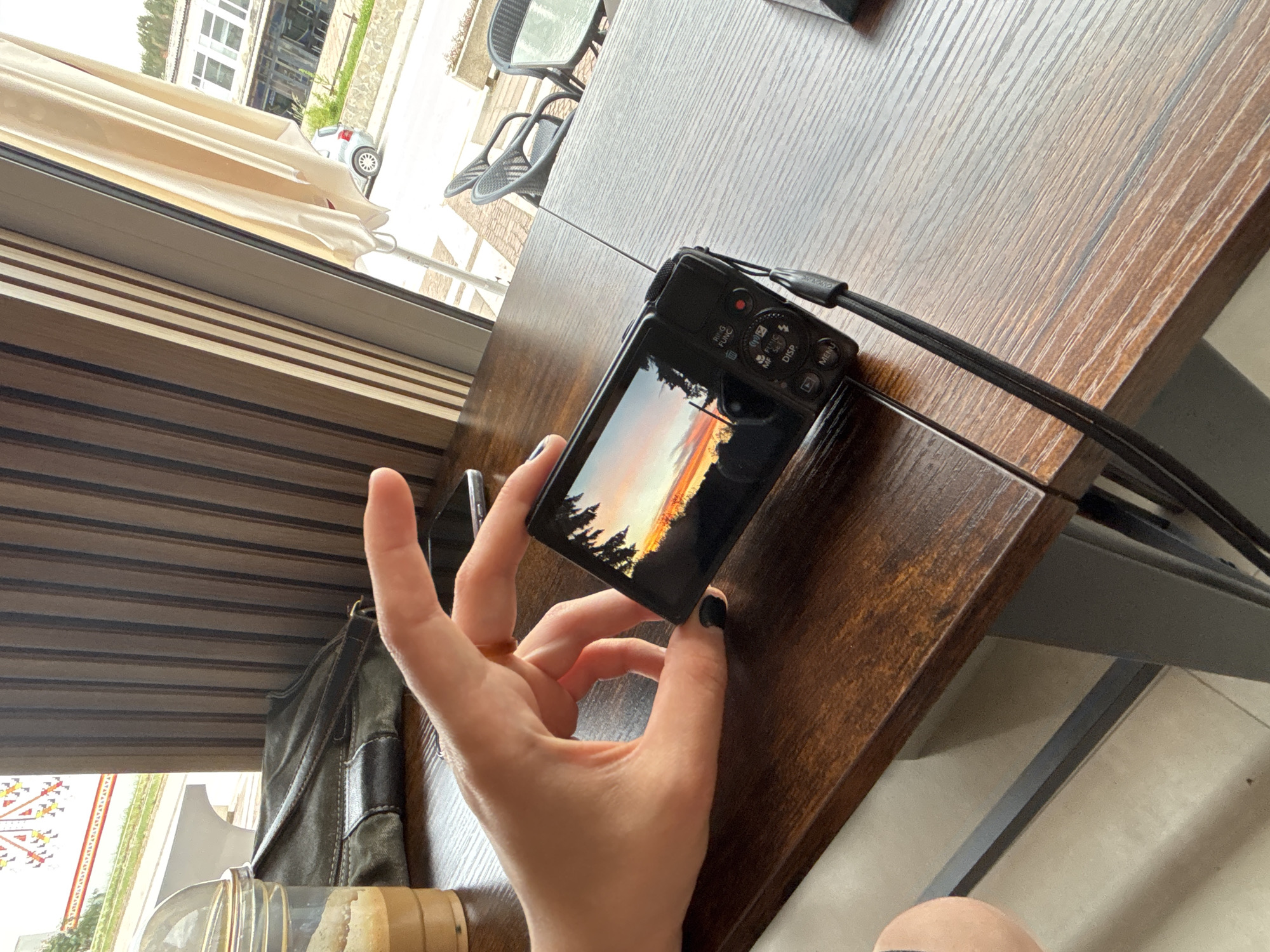 A close-up, angled, indoor shot shows a person holding a black digital camera displaying a landscape photo of trees against a sunset, likely near Garmen, Bulgaria. The camera is positioned above a brown wooden table. The person's hand, adorned with a ring on the ring finger and black nail polish, is holding the camera. A window is visible in the background, showing an outdoor scene with buildings, possibly a cafe or restaurant setting. The environment suggests a relaxed, daytime setting.