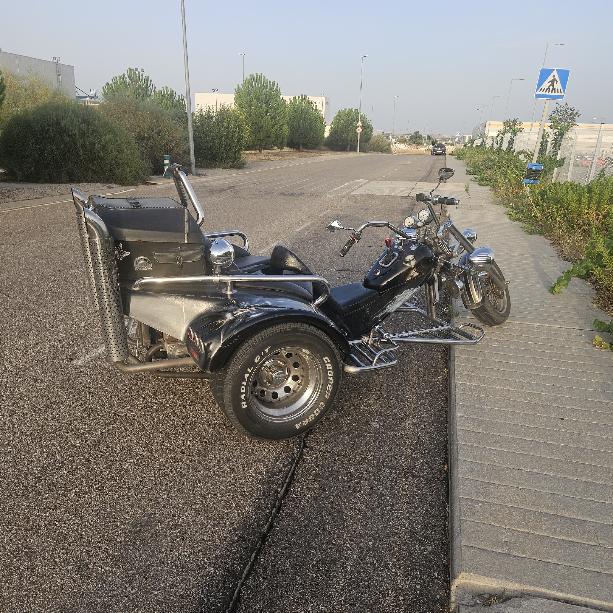 The image shows a black custom trike parked on the side of a road next to a paved walkway. The trike has a large, studded exhaust pipe on the left rear side and a black leather seat and storage compartment. It is positioned with its front wheel on the road and its two rear wheels on the concrete walkway. The background includes a street with lane markings, industrial buildings, trees, and streetlights. A pedestrian crossing sign is visible in the distance on the right. The scene appears to be in an industrial or commercial area of Illescas, Spain, likely during daylight hours with clear skies.