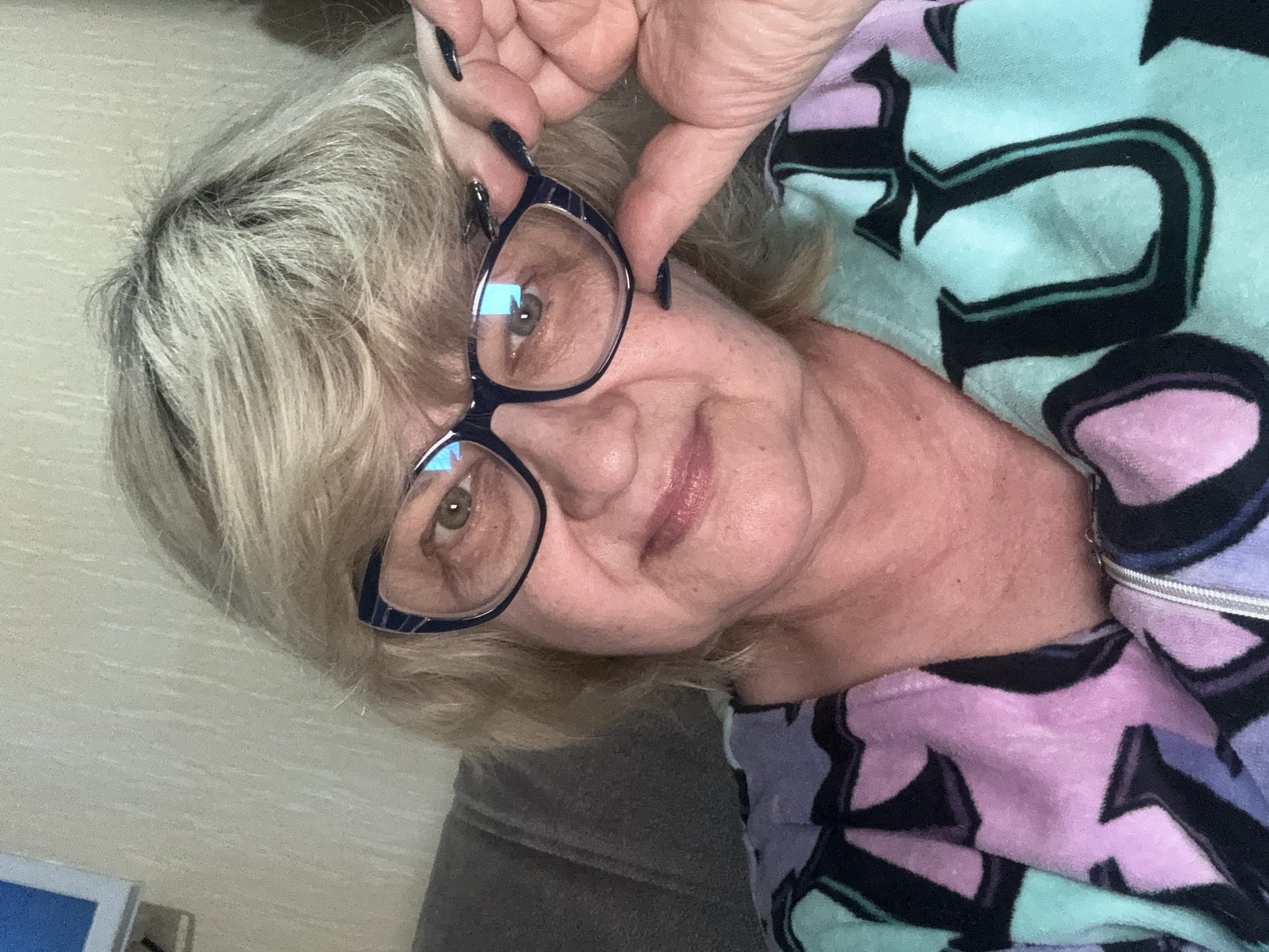 A middle-aged woman with blonde hair and glasses is taking a selfie indoors. She is wearing a colorful pajama top with a zippered neckline and has dark nail polish on her fingers. The background appears to be a wall with a textured pattern and a piece of furniture, possibly a sofa, is visible to the left. The lighting suggests an indoor environment, likely a home setting in Mytishchi, Russia.