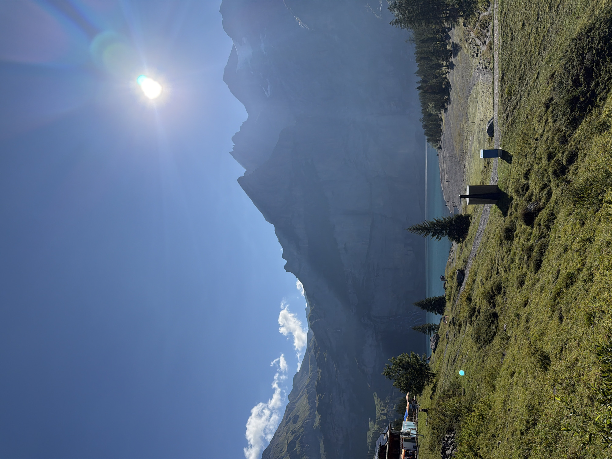 The image depicts a serene mountain landscape in Kandersteg, Switzerland. In the foreground, a grassy slope descends towards a clear turquoise lake. A few scattered trees dot the landscape, and a path winds its way down towards the lake.
On the left side of the image, a building complex is visible, with umbrellas providing shade for outdoor seating. Further down the slope, near the path, are two structures that appear to be public restrooms or utility boxes.
The middle ground is dominated by the tranquil lake, with a stony shore where a few individuals can be seen walking. A dense forest lines the far bank of the lake, with tall evergreen trees.
In the background, majestic, rugged mountains rise steeply, their peaks partially shrouded in mist and haze. Sunlight streams down from the upper right corner of the frame, creating a lens flare and casting long shadows across the scene. The sky is a vibrant, clear blue, with a few wispy clouds present. The overall atmosphere is peaceful and natural.