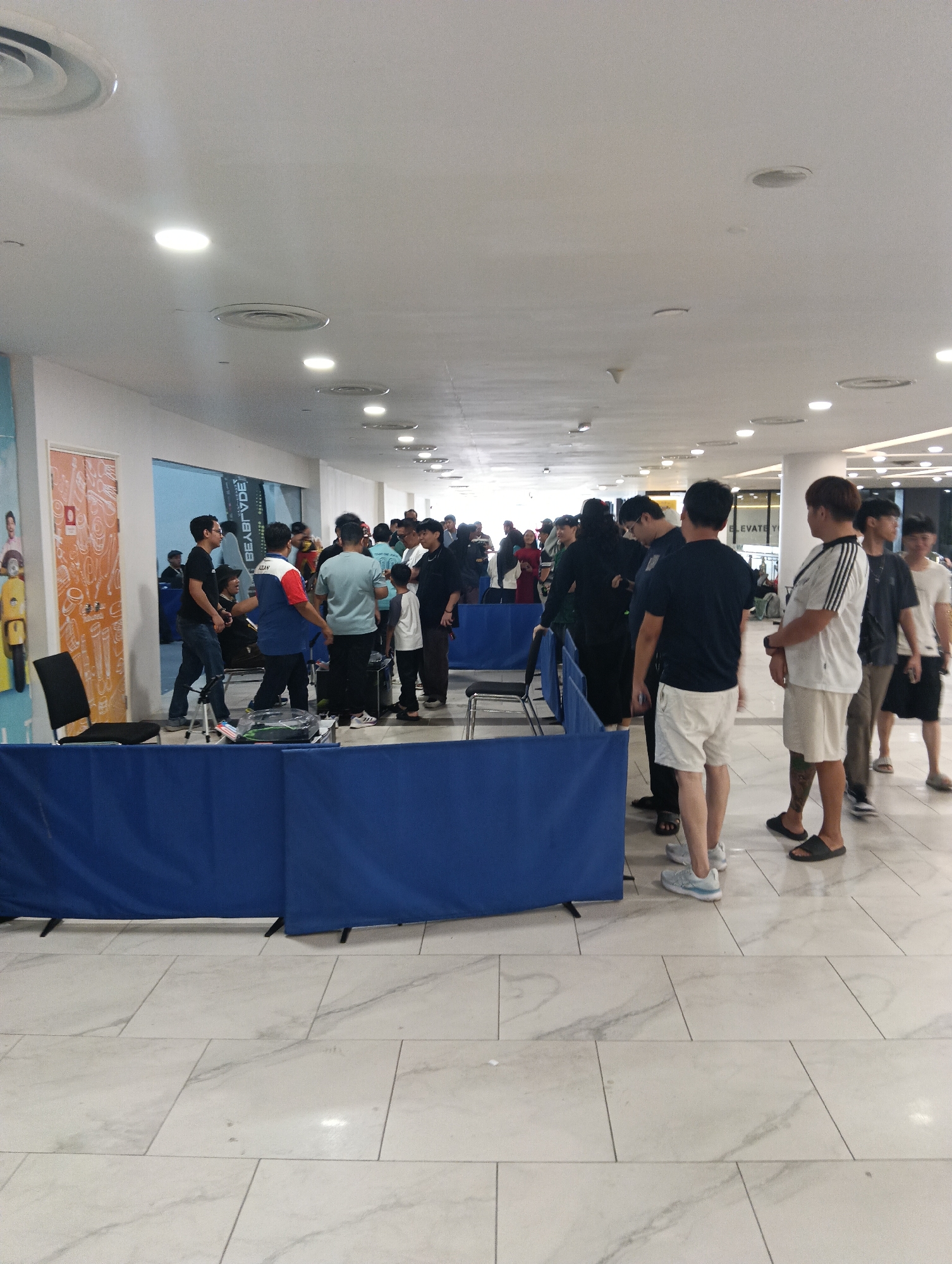 The image depicts an indoor environment, possibly a convention or exhibition hall, with a crowd of people gathered. The scene appears to be set up for an event, indicated by blue fabric draped across tables and chairs arranged in rows. Several individuals are standing and interacting, some appearing to be participants or attendees. The flooring is tiled with a light marble pattern. The ceiling features recessed lighting and circular air vents. In the background, there are walls with posters and some signage. The overall impression is a busy, organized gathering. Based on the visual elements, it's difficult to pinpoint the exact location within Kuching, Malaysia, without additional context. However, the environment suggests a public venue such as a shopping mall, exhibition center, or community hall that hosts events.