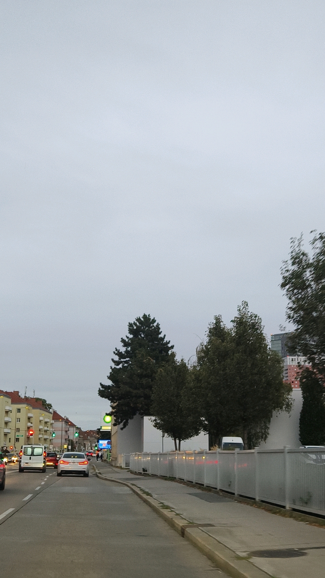 The image depicts a street scene in Vienna, Austria, under an overcast sky. The perspective is from a vehicle moving along a multi-lane road. On the left side of the road, there are apartment buildings with yellow facades and traffic lights indicating red and green signals. Several cars are visible on the road, including a white van and a white sedan. On the right side of the road, there is a sidewalk bordered by a white fence. Behind the fence, there are several trees, some of which are tall evergreens. Further in the background, a modern building with a glass facade and some red-patterned elements is visible, along with other city buildings. A BP gas station sign is also present. The overall atmosphere is urban and subdued, likely due to the weather.