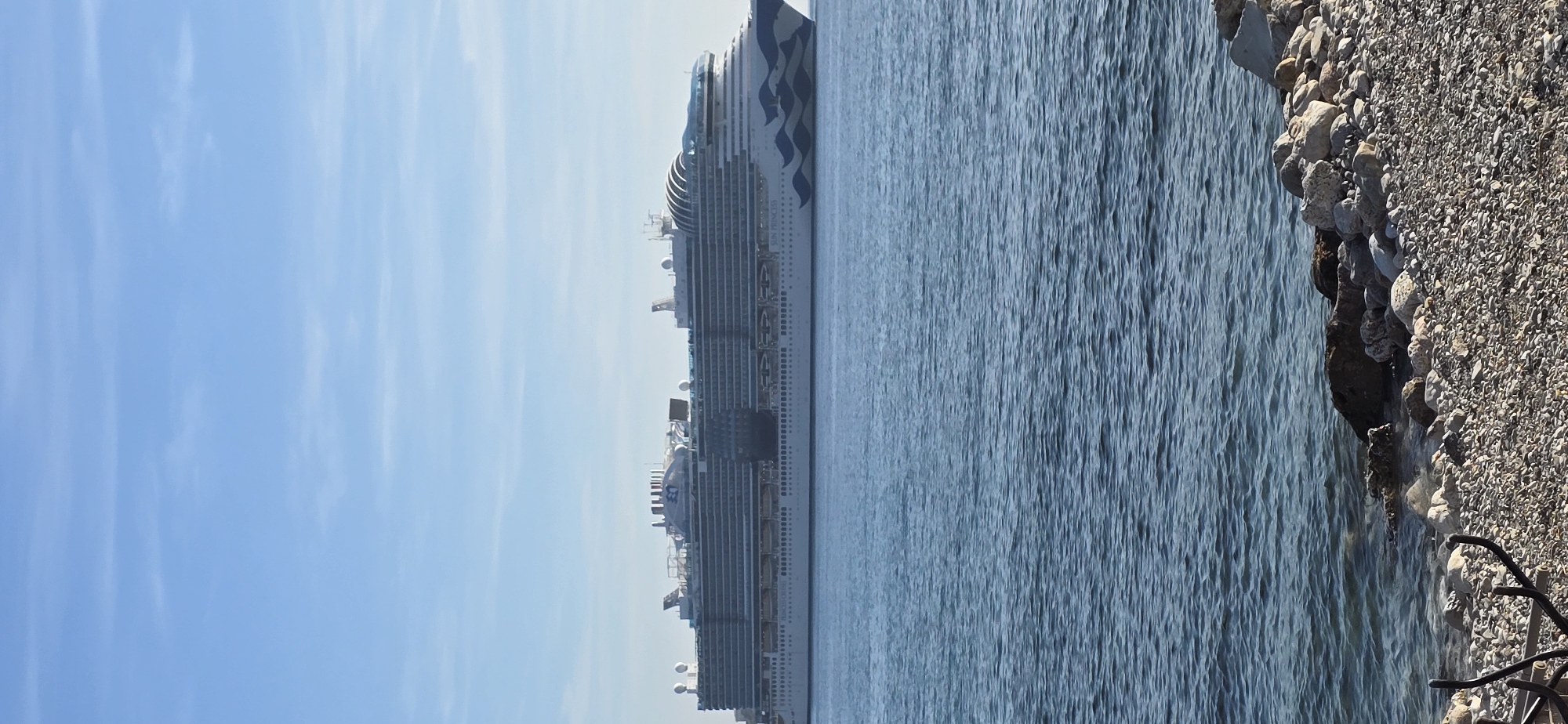 A large cruise ship is prominently featured in a bright daytime coastal scene, likely in Bar, Montenegro. The image is composed with a clear blue sky filling the left portion, the cruise ship positioned horizontally across the middle on the water, and a rocky, pebbly shore on the right.
The sky is a serene light blue with a few wispy, barely visible clouds. The water, a calm blue-grey, shows subtle ripples and reflections, indicating still conditions. The cruise ship is a massive vessel with multiple decks, balconies, and lifeboats lining its sides. Its bow features a distinctive dark blue wave-like design against the white hull. Near the stern, the word "PRINCESS" is clearly visible on the ship's side, identifying it as a Princess Cruises vessel. A large Princess Cruises logo, a stylized sea witch or mermaid with a crown, is also discernible. Various radar domes and antennae are scattered on the upper decks of the ship.
The shore on the right consists of a mix of larger, weathered rocks and smaller pebbles, ranging in color from light grey to dark brown. Towards the very bottom right, some dark, tangled debris or a natural root structure is partially visible on the shore. The entire scene is bathed in bright daylight, suggesting fair weather and good visibility. The ship appears to be stationary, either docked or anchored close to the land.