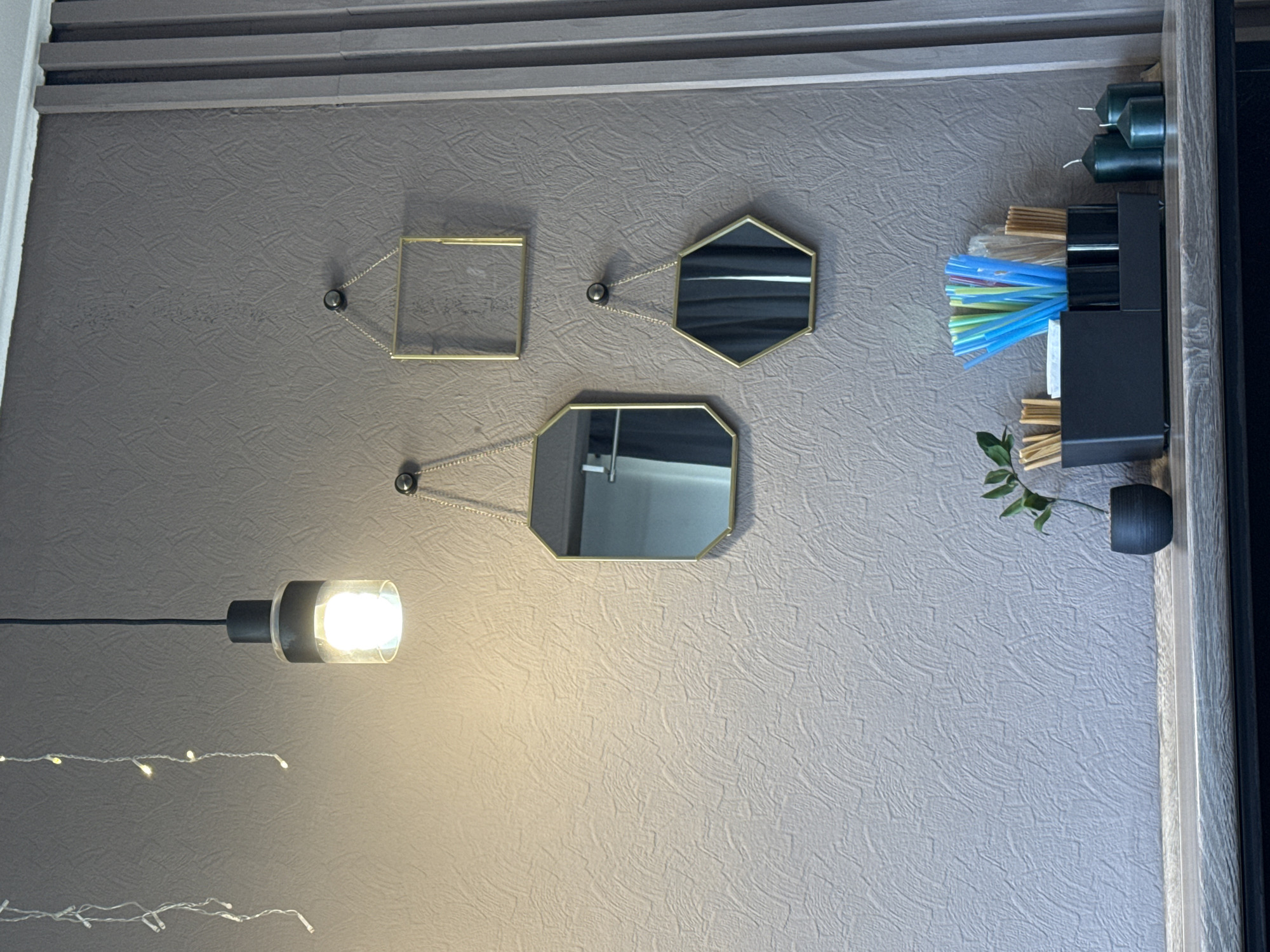 The media file presents an indoor scene showcasing a textured, greyish-brown wall, possibly from Chaikovsky, Russia. At the top, horizontal grey panels with distinct ridges are visible, suggesting part of a ceiling or upper wall structure.
Three decorative mirrors with golden frames are mounted on the wall, each suspended by small, delicate chains attached to dark, circular fasteners. One mirror is square, while the other two are octagonal, arranged in a vertical configuration below the square one. The mirrors reflect indistinct portions of the room, showing variations in light and dark tones.
A hanging light fixture with a clear, cylindrical bulb and a dark base is positioned on the left side, casting light onto the lower section of the wall. Below this light, two strands of fairy lights, loosely draped horizontally, add to the decorative ambiance.
On the right, a vertical shelf unit with a wooden-look frame holds several objects. The top shelf contains multiple dark green, stubby candles. Below this, a black compartmentalized organizer is visible. Its left section is filled with bundles of light brown sticks, potentially incense or matches. The central compartment overflows with a collection of colorful plastic straws, primarily blue, yellow, and green. The rightmost section contains white block-shaped items. A small dark pot with a green leafy plant is situated on a lower shelf, peeking into the frame.
The image captures a static interior setting without any human presence or ongoing activity. The artificial lighting from the hanging bulb suggests an indoor environment where natural light might be limited, or it could be evening. No discernible text is visible anywhere in the frame. The overall impression is one of an organized and thoughtfully decorated space.