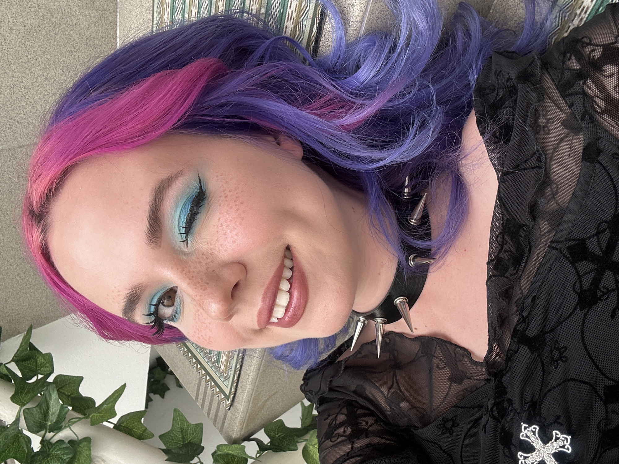 A young woman is featured in a rotated close-up portrait, appearing to lie on her side and smiling warmly at the viewer. Her hair is a striking split dye, with vibrant pink on one side and flowing purple and lavender strands on the other. She has fair skin, with visible freckles across her nose and cheeks. Her eye makeup is prominent, featuring bright blue eyeshadow with sharp winged black eyeliner and dark mascara. She wears a black choker adorned with several silver spikes and a black top or dress crafted from sheer, intricately patterned mesh fabric, possibly with velvet flocking details. A silver, jewel-encrusted cross pendant rests on her chest. In the foreground, artificial green ivy leaves are decoratively placed. The background includes a textured grey surface and a patterned textile, suggesting an indoor setting in the City of Zagreb, Croatia. She conveys a happy and friendly emotion with her wide smile.