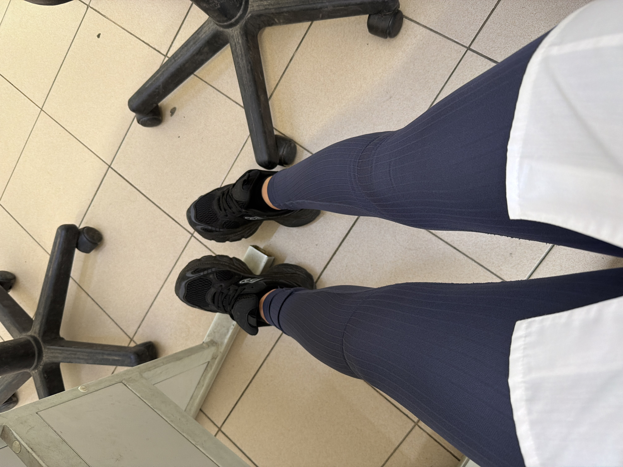 The image presents an overhead view of a person's lower body within an indoor setting in Sofia, Bulgaria. The subject is wearing dark blue, form-fitting leggings with a ribbed texture, paired with black athletic shoes.
The setting features a light-colored tiled floor with dark grey grout lines, creating a grid pattern. Several objects are visible on the floor, including the black, multi-pronged bases with wheels of what appear to be at least two office chairs. On the lower left and extending towards the center, a light-colored, possibly metallic, structure resembling part of a table or bench is also present.
The perspective is directly looking down, capturing the legs and the floor immediately around them. White fabric, likely part of a garment such as a lab coat or uniform, is visible on both sides of the legs, suggesting a professional or institutional environment like a clinic, hospital, or office. No specific activity is depicted beyond the person being present in this space. There are no discernible emotions, time of day cues, or visible text within the frame.