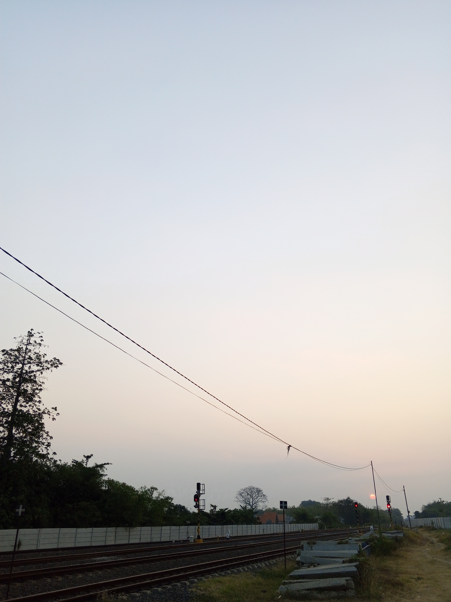 The image captures a railway track scene at dusk in Pakusamben, Indonesia. Two sets of parallel railway tracks dominate the foreground, with ballast and gravel filling the space between them. A grey concrete wall runs along the left side of the tracks, separating them from a dense line of green trees. Several electrical wires are strung across the upper portion of the frame, supported by poles.
In the mid-ground, a railway signal with a red light is visible on the right side, indicating a stop. Further to the right, a faint red circle, likely the setting sun, is visible low in the hazy sky. Bare trees and more foliage are scattered throughout the landscape. The sky transitions from a pale blue at the top to a soft orange and yellow near the horizon, suggesting the end of daylight. On the left side of the image, a single, tall, leafless tree stands silhouetted against the sky. The ground next to the tracks on the right is covered in dry grass and dirt, with some concrete blocks piled up. No people are visible in the frame.