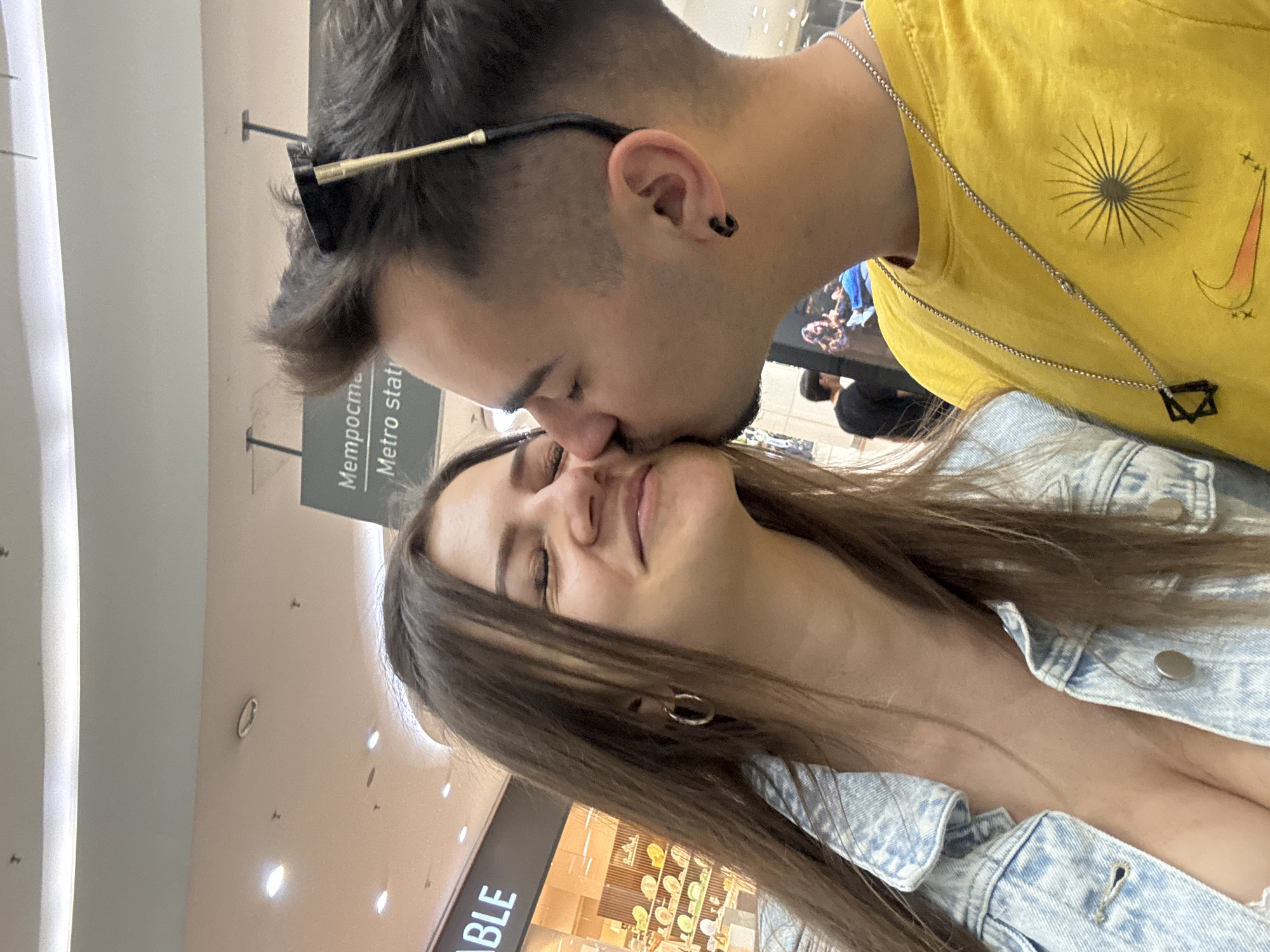 A couple poses for a photo indoors. The man is kissing the woman's nose. He wears a yellow shirt with a sunburst design, a silver chain necklace with a Star of David pendant, and sunglasses perched atop his head. The woman is wearing a light blue denim jacket. They appear to be in a shopping center. The background includes signs, lighting fixtures, and storefronts. A sign with Cyrillic script including the words "Metro statio" indicates the location as likely being near a metro station in Sofia, Bulgaria.
